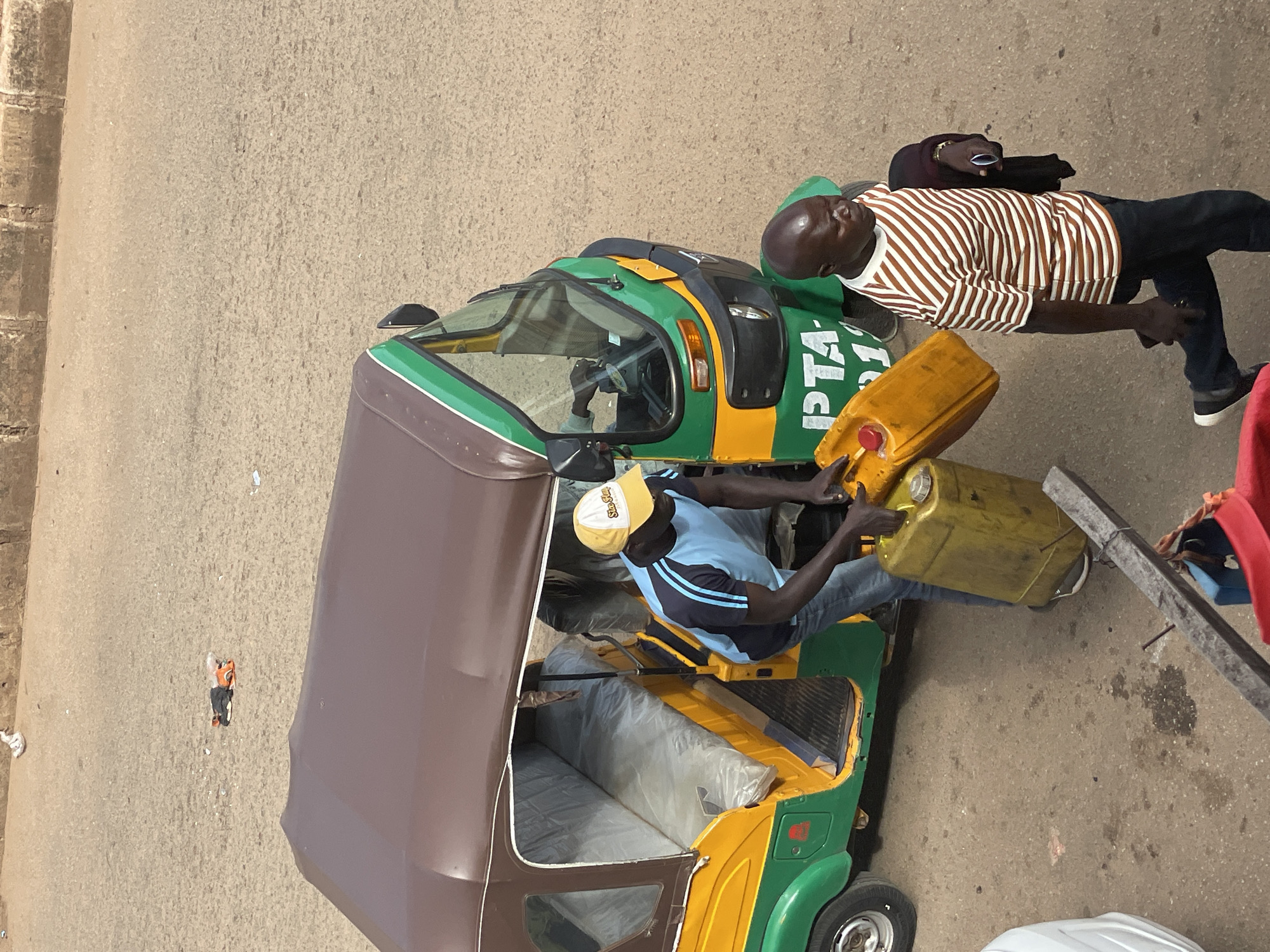 In the city of Unknown, Unknown, two men are interacting near a green and yellow auto-rickshaw. One man, wearing a blue and black striped shirt, blue jeans, and a yellow and white baseball cap, is holding a large yellow fuel can. He appears to be either filling or emptying the can into the auto-rickshaw. The auto-rickshaw has a brown roof and is parked on a paved surface. The other man, wearing a brown and white striped t-shirt and dark pants, is walking away from the auto-rickshaw. He is carrying a dark-colored garment over his left shoulder and holding a smartphone in his right hand. In the background, there is a rough concrete wall and a dusty, sandy ground. A pile of clothes, including orange and black items, is visible on the ground in the distance.