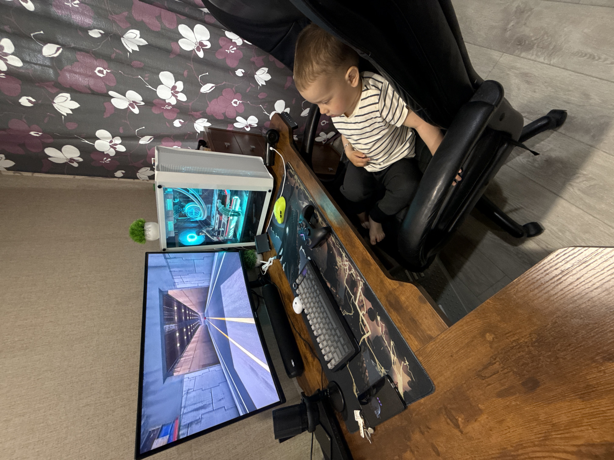 A child is sitting in a black office chair at a desk with a computer, keyboard, mouse, and monitor, presumably in a residential setting in Rusanovo, Russia. The monitor displays an image of a tunnel. The desk is wooden and covered with a mousepad. A computer tower with internal lights is on the desk next to the monitor. A small potted plant is near the computer tower. The wallpaper features a floral pattern. A set of keys and a smartphone are on the right side of the desk.