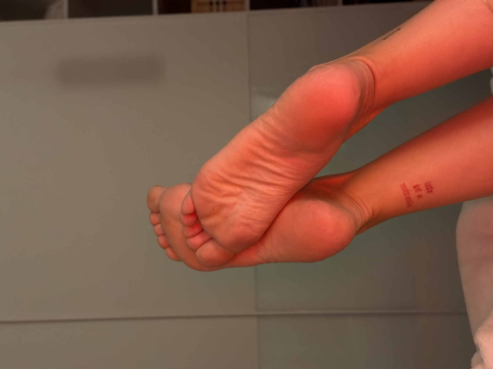 A close-up, horizontally-oriented shot shows a pair of bare feet crossed at the ankles, with the soles facing the viewer. A red tattoo is visible on the inner part of one ankle. The background appears to be a neutral-colored surface with a cabinet. Location information is not available.