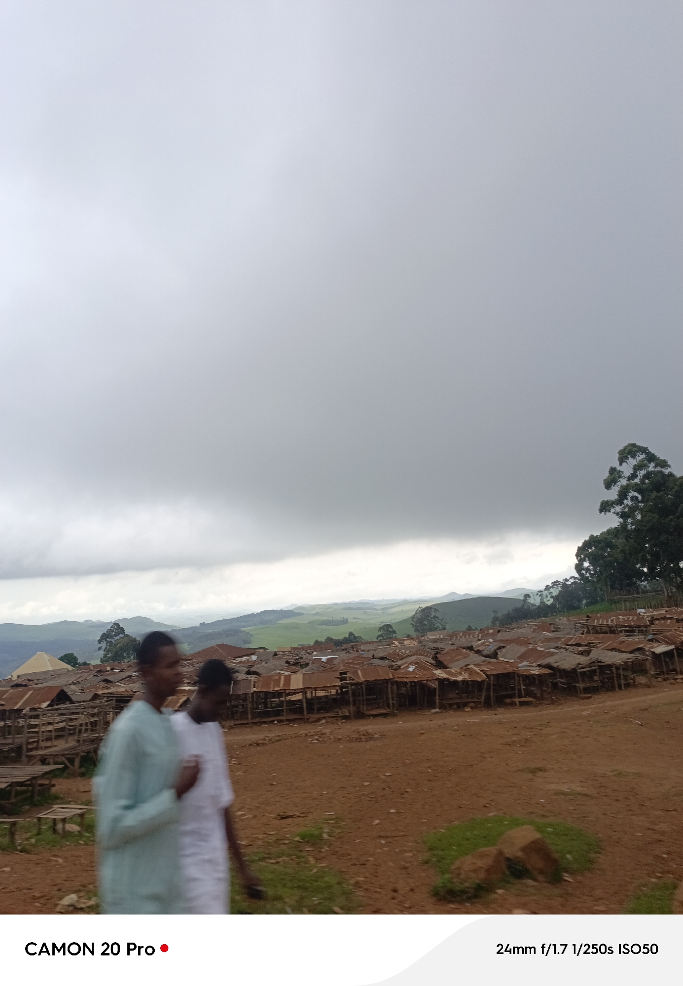 The image shows two individuals walking in a dusty area in front of a settlement with numerous structures featuring corrugated tin roofs. The settlement appears to be located on a hillside, with rolling green hills and trees visible in the background under a cloudy sky. The overall scene suggests a rural or semi-urban environment in the city of Nguroje, Nigeria.