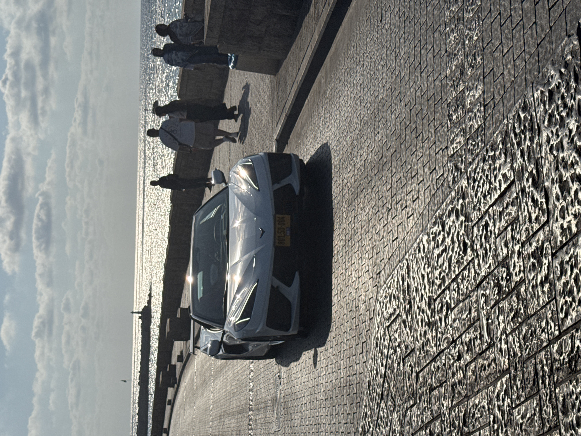 The image captures a bright, sunny day at the Tel Aviv promenade, likely near the Jaffa port or a similar coastal area. The foreground features a cobblestone pavement that is partially wet and reflects the sunlight, creating a shimmering effect. A white convertible sports car, possibly a Chevrolet Corvette C8, is parked on the right side of the road, facing slightly away from the camera. Its headlights are illuminated, and a license plate that reads "801-50-004" is visible.
In the middle ground, several people are walking along the promenade, silhouetted against the bright sea. They appear to be enjoying the view and the weather. Some are walking in groups, while others are alone. The sea itself is a brilliant blue, with sunlight glinting off its surface. A long, stone breakwater extends into the sea, with a small lighthouse visible at its end. The sky is partly cloudy, with fluffy white clouds scattered across a blue expanse. The overall impression is one of a pleasant day by the sea in a vibrant city.