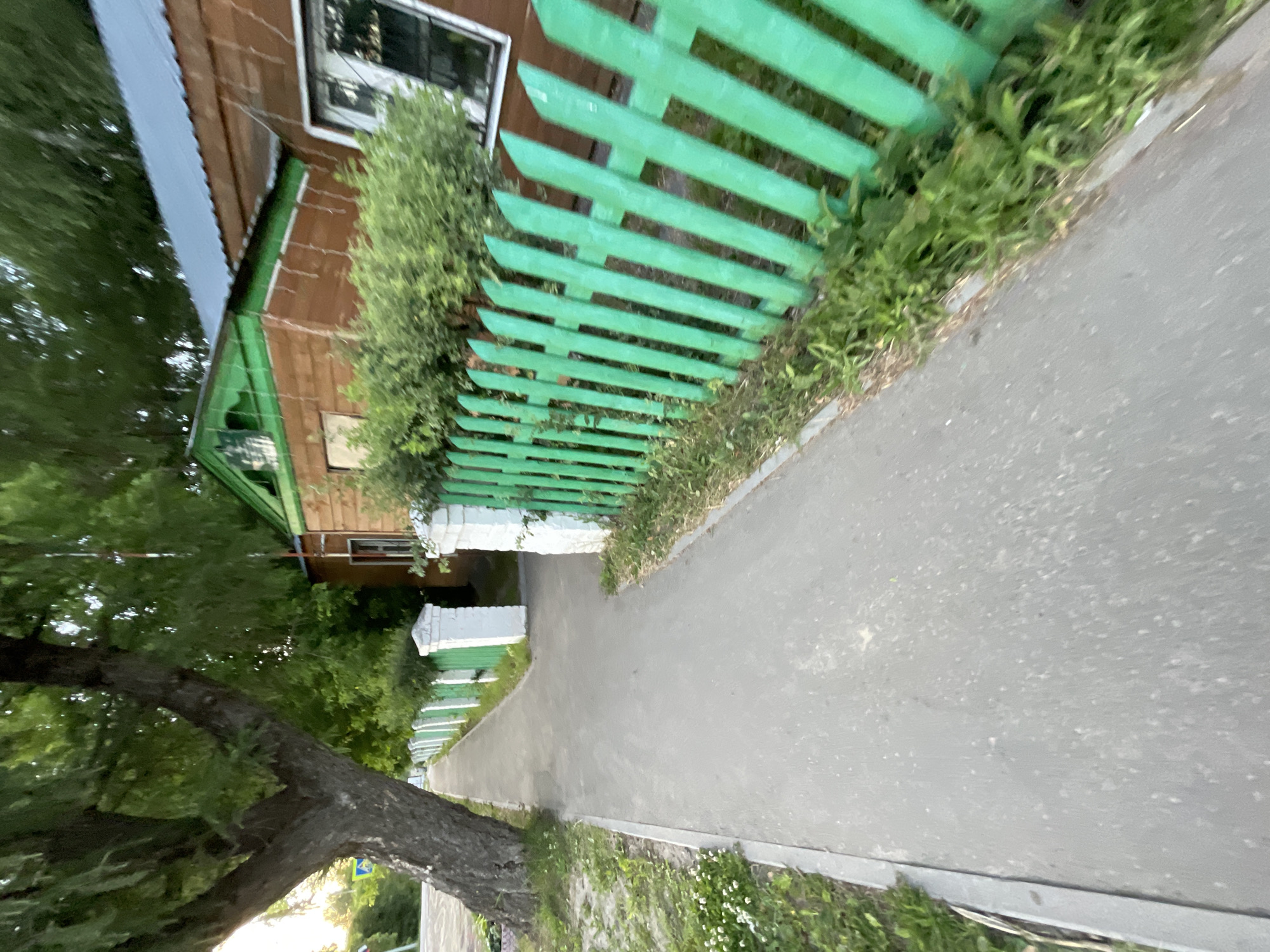 The image shows a sidewalk next to a wooden house with a green fence in Krasnye Baki, Russia. The house is made of brown wood and has a metal roof. The fence is painted green and has white pillars. There is a large tree on the left side of the image, and green bushes are planted behind the fence. The sidewalk is made of asphalt, and there is grass growing on the side of the sidewalk. The scene appears to be a residential area in a town or village.