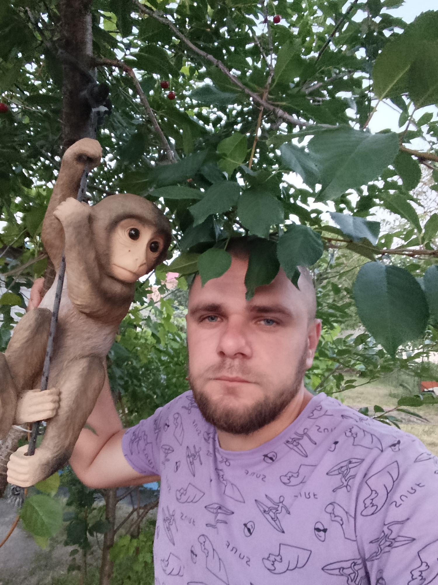 A man with short hair and a beard, wearing a purple t-shirt with a repeating pattern of cartoon surfers, is posing for a selfie outdoors. He is to the right of a life-sized statue of a monkey hanging from a branch. The background consists of green leaves, branches, and some red cherries on a tree. The image appears to have been taken during daylight. The man is looking directly at the camera with a neutral expression. The monkey statue is positioned to the man's left, with its left arm draped over a branch and its right hand appearing to grip a thin rope or wire. The environment suggests a garden or a backyard setting.