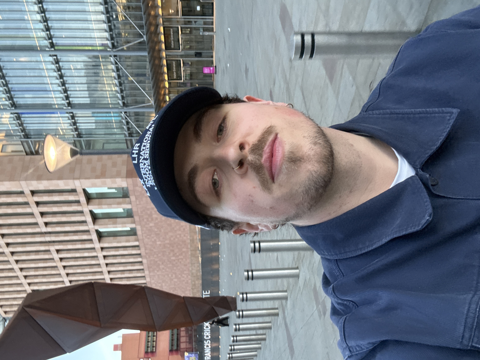A medium shot depicts a light-skinned male wearing a dark blue baseball cap emblazoned with "LHR International Room Service" and a dark blue button-down shirt. He has light facial hair, a nose ring, and an earring. He is looking directly at the camera.
The background features modern architecture including a building with multiple glass windows and exposed metal beams and a sculpture consisting of many geometric metal planes in bronze or brown color. A row of metallic poles line the foreground. The word "FRANCIS CRICK" is partially visible.
The location is the Francis Crick Institute in London, United Kingdom.