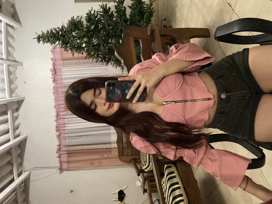 A young woman with long, dark hair takes a selfie indoors. She wears a pink, short-sleeved crop top with a zipper and dark shorts. The background includes a window with white trim, a light pink curtain, and a green artificial Christmas tree. Parts of wooden furniture and a black office chair are also visible. The woman holds a smartphone with a colorful case up to her face.