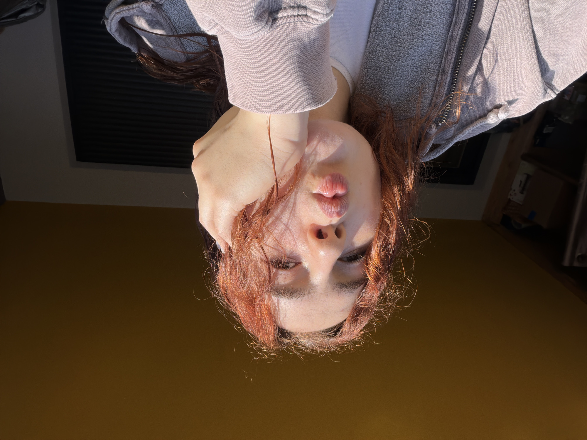 A woman with long, reddish-brown hair is seen from a low angle, with her head tilted upside down. She is wearing a gray hoodie. Her left hand is raised to her face, partially obscuring it. The background consists of a yellow surface, a black rectangular object (likely a window or panel), and a partial view of shelves and cabinets. There is no indication this image was taken in Strasbourg, France.