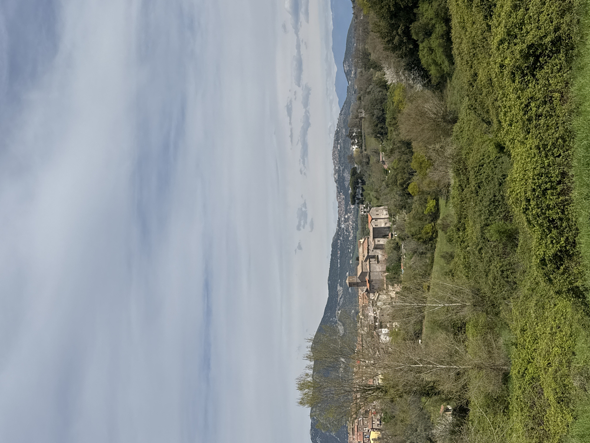 The image depicts Rignano Flaminio, Italy. The scene includes a mountain range in the distance, with green, lush vegetation covering the landscape to the right. A small town with buildings featuring red-tiled roofs can be seen in the center. A blue sky with white clouds occupies the upper left portion of the image. A few bare trees are visible in the foreground.