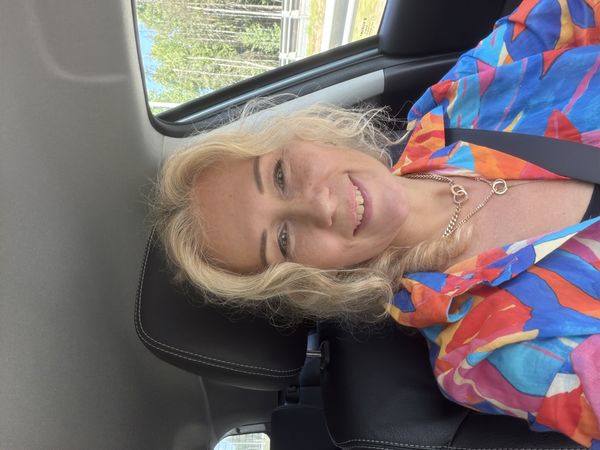 The media file is a close-up selfie of a woman taken inside a car. The woman has blonde, wavy hair and is smiling at the camera. She is wearing a colorful, abstract-patterned shirt with shades of blue, orange, pink, and yellow. A gold necklace with two interlinked rings is visible around her neck. The background shows the interior of a car, with black leather seats and a portion of the car's interior ceiling. Through the car window, a glimpse of trees and a blue sky can be seen, suggesting an outdoor setting and daytime. There is also some text visible on the window, which appears to be "SUZUKI" and "E1" which are likely car manufacturer and model related. The overall mood is cheerful and casual. The location context provided is Rokhma, Russia.