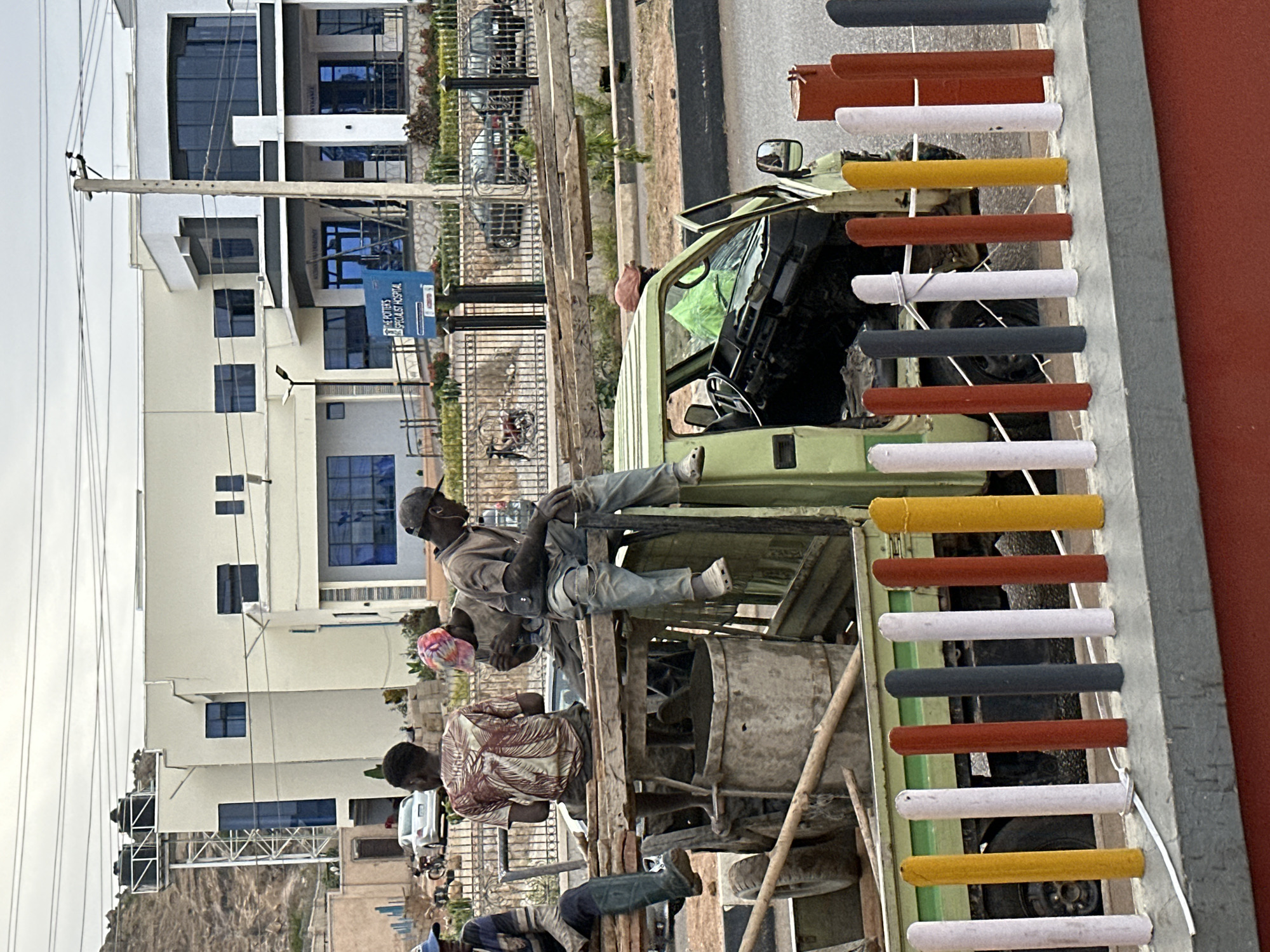 This image, capturing a street scene in Jos, Nigeria, depicts several men and a light green pickup truck under an overcast or hazy daytime sky.
In the foreground, a light green utility pickup truck is central, its open bed filled with large metal drums or containers, possibly for mixing concrete, and various wooden poles and tools. One man, wearing a cap, light-colored shirt, and jeans, is seated on the left edge of the truck bed, looking to his right. Another man, wearing a distinct patterned shirt, stands within the truck bed to the lower left. A third individual is partially visible at the bottom left, also within the truck bed. A fourth man, wearing an orange hat, is seen near the cab of the truck in the middle ground. Their presence and the equipment suggest manual labor, likely related to construction or transport.
To the right of the truck, a vibrant barrier or fence stands out. It comprises a series of alternating white, yellow, orange-brown, and dark grey cylindrical elements mounted horizontally on a grey rail, positioned against a solid red wall.
In the background, a multi-story building with a light-colored facade and dark blue tinted windows dominates the left side. A blue sign in front of this building clearly displays the text "THE POTTER'S SPECIALIST HOSPITAL" and "ENTRANCE" at the bottom, with additional text below "HOSPITAL" that is partially obscured but appears to include "NURSING". Further into the background, other vehicles are parked behind a metal fence, and utility poles with power lines crisscross the upper frame. The overall setting is an urban or semi-urban environment, showing a typical working day.