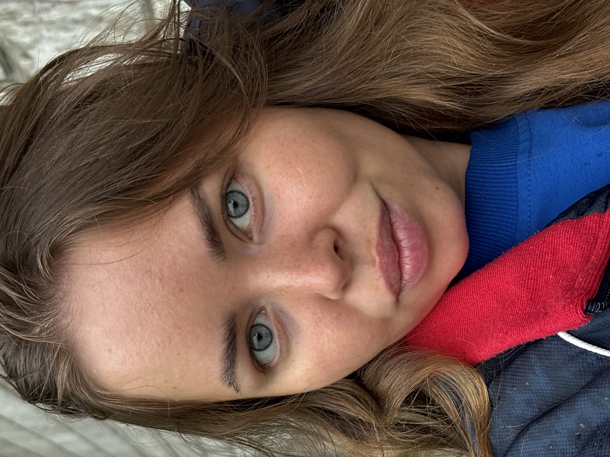 A close-up, slightly rotated, portrait photograph depicts a fair-skinned woman with light brown, wavy hair. Her hair frames her face and extends beyond the shot. She has light blue eyes and appears to have minimal or no makeup, with a slight smile. Freckles are visible on her face. She is wearing a blue top with a red portion near her neck. The image's background is nondescript. There is no information in the image to connect it to the city of Русский Вожой, Russia.