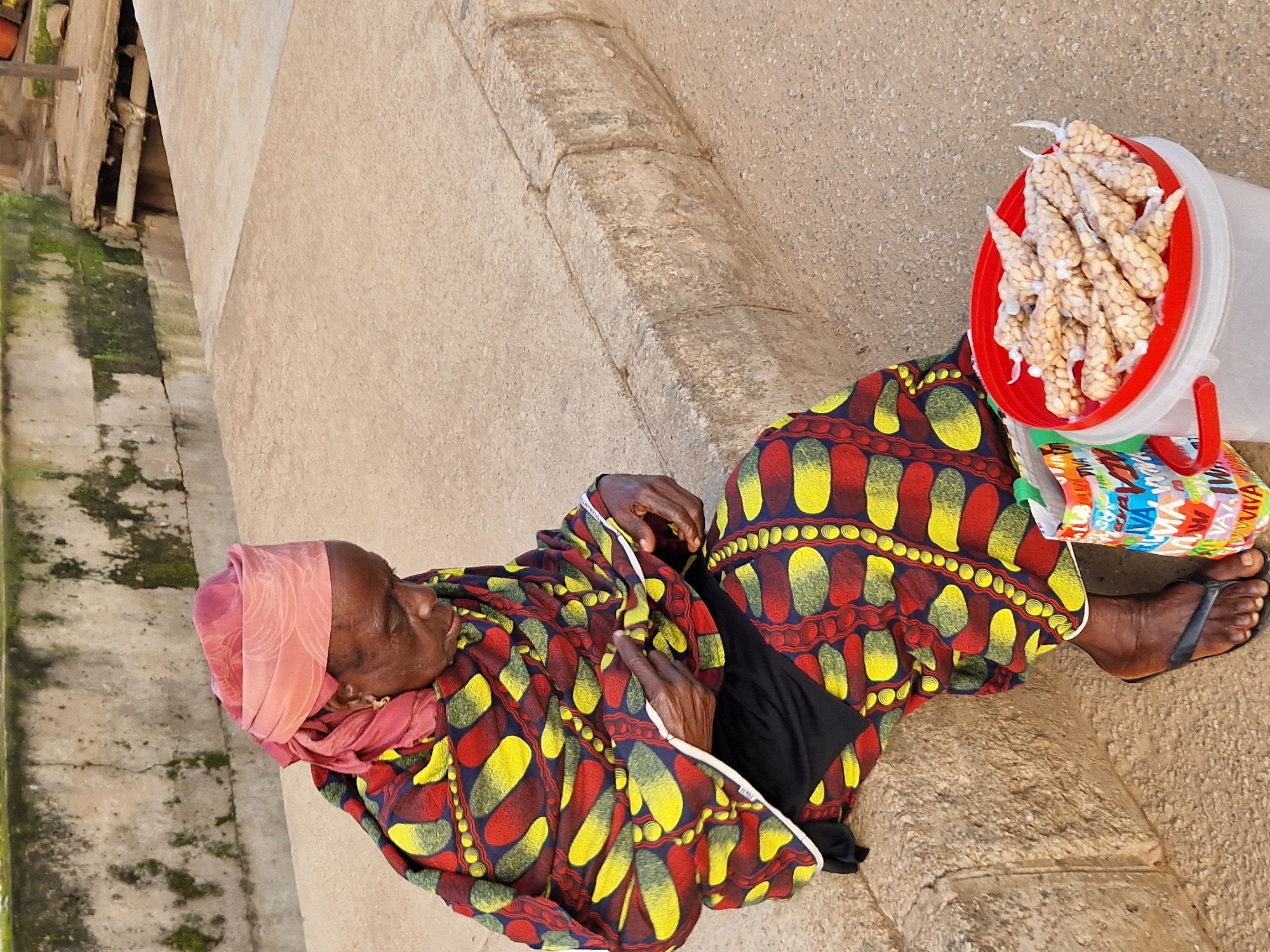 In Kabong, Nigeria, an elderly woman wearing a colorful, patterned wrap dress and a pink headscarf is seated on a concrete curb. She is looking down at her hands, which are positioned in her lap. Beside her, on the ground, is a red-rimmed white bucket filled with small, white, bean-like objects wrapped in clear plastic. A multi-colored patterned shopping bag rests next to the bucket. The background consists of a weathered concrete wall with green mossy patches and a sandy, paved surface.