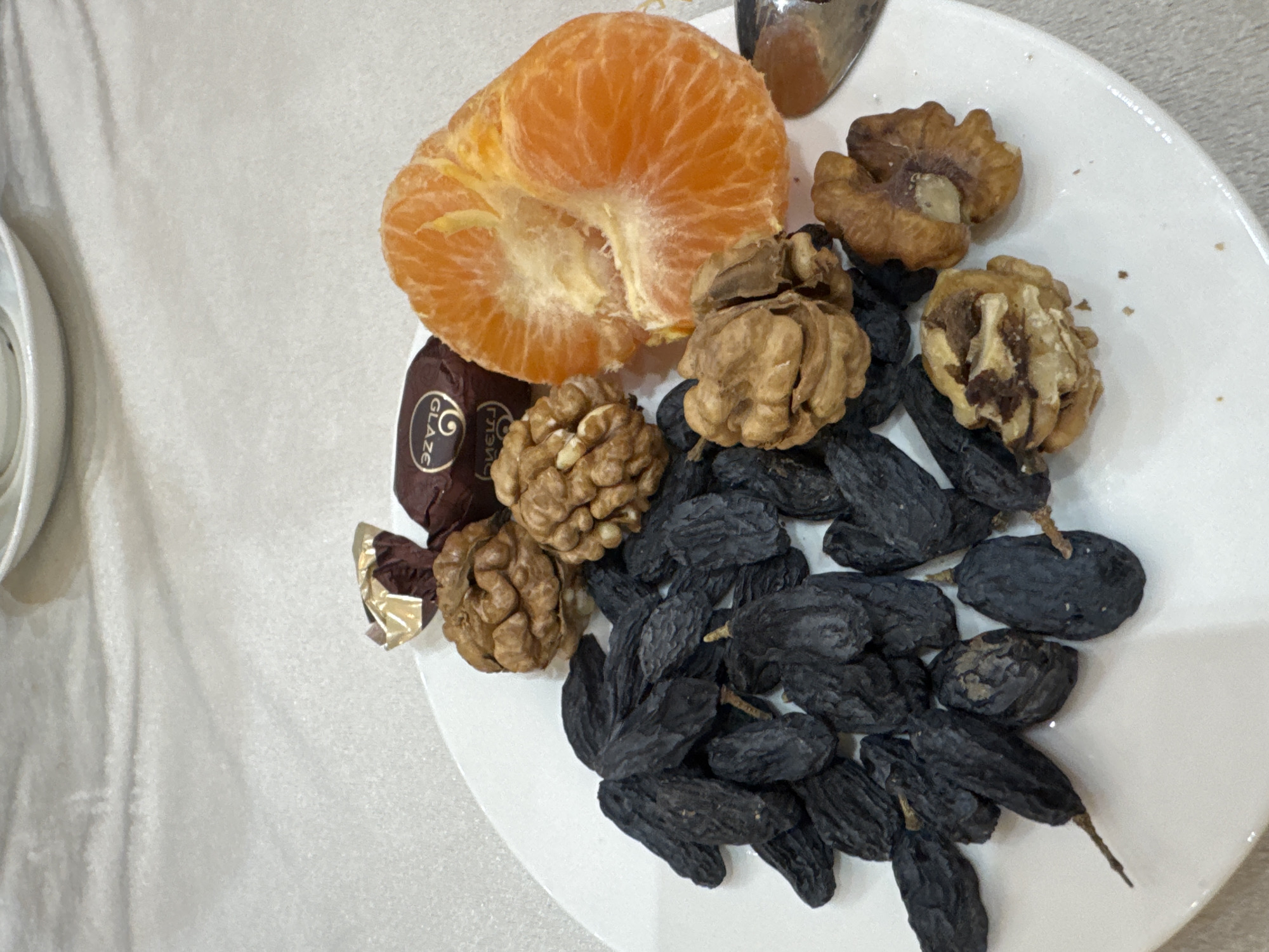 A white plate is arranged with food items: an open tangerine, several walnuts (some open), a pile of dark raisins, and a wrapped candy. The candy wrapper has the word "GLAZE" and some Cyrillic text. The scene is on a surface with a slightly textured, off-white color. It's likely an indoor setting. There's no direct evidence linking this scene to Samarkand City, Uzbekistan; that connection is speculative based on the user's prompt.