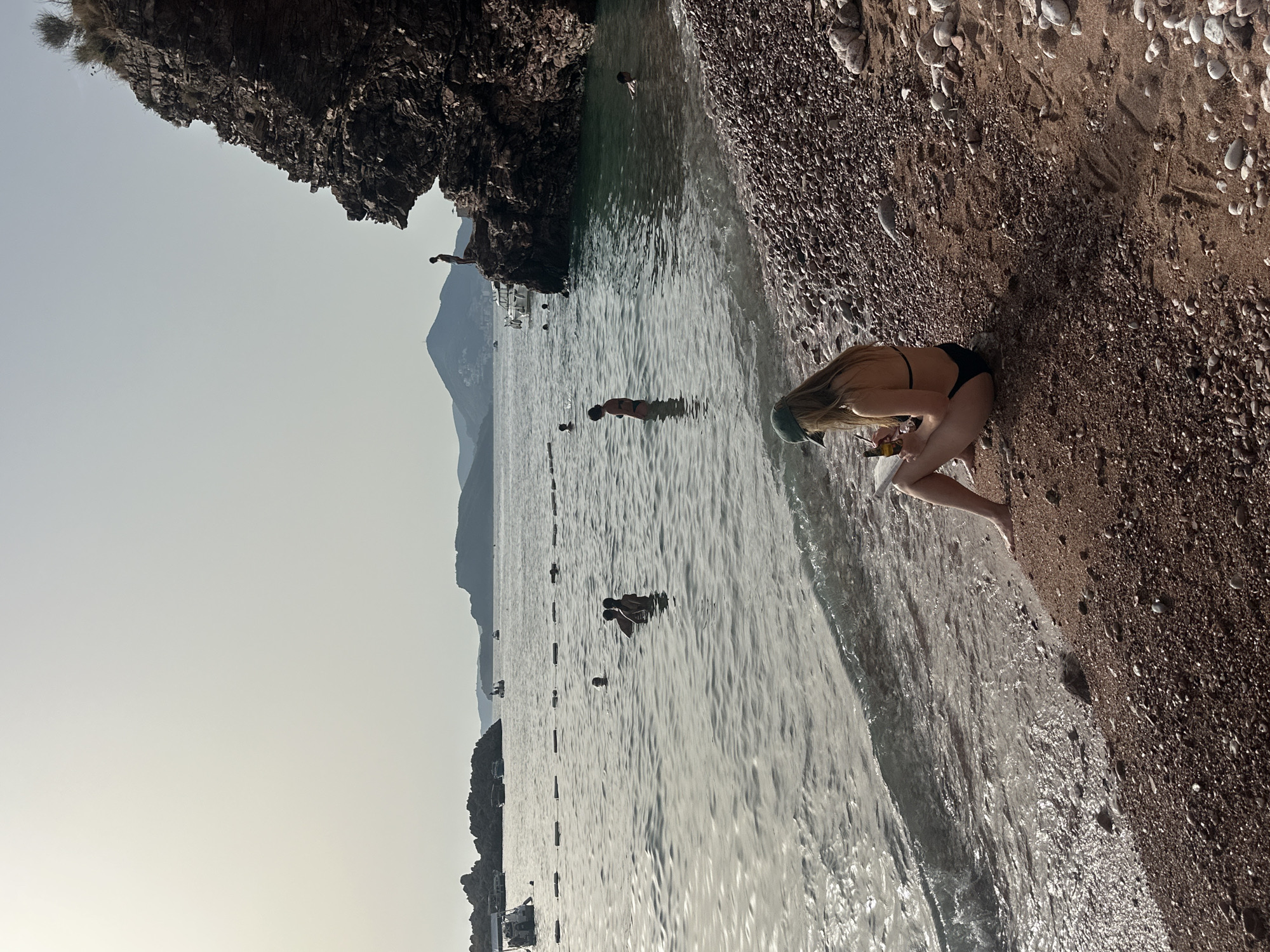The image depicts a sunny day at the beach in Sveti Stefan, Montenegro. In the foreground, a woman with long blonde hair, wearing a green cap and a black bikini, is sitting on the pebble beach, facing the sea. She is holding a book and a bottle. Her legs are bent, and her bare feet are near the water's edge.
The sea is calm with gentle waves lapping at the shore. There are several people swimming and wading in the water. In the middle ground, a group of people are visible in the sea, some of them are close together. Further out, a bouy line with floats marks a swimming area.
To the right, a rocky cliff juts out into the sea, with a person standing on its edge, silhouetted against the sky. Boats are anchored in the distance. The background shows a hazy coastline with hills and mountains, characteristic of the Montenegrin coast. The overall scene is one of leisure and relaxation by the Adriatic Sea.