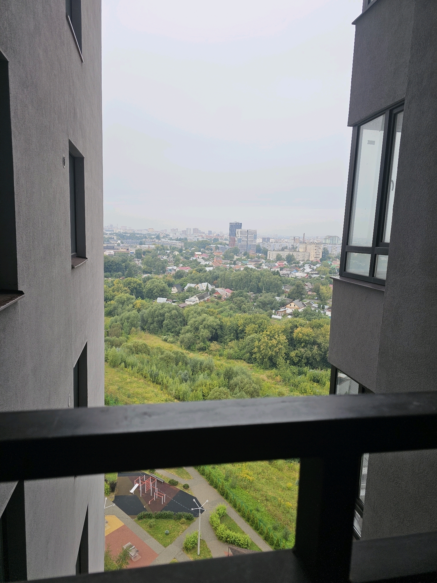 The image shows a view from a high-rise building overlooking a city, likely in Ryazan, Russia. The foreground is dominated by a dark railing. Through the railing, a playground with red and white equipment is visible on the ground level, adjacent to a paved pathway and a grassy area. Beyond the playground, a dense green forest stretches towards a residential area with houses. In the distance, a cityscape with various buildings, including a modern, tall, multi-story structure, is visible under an overcast sky. The scene appears to be an urban environment with a mix of residential, natural, and constructed elements.