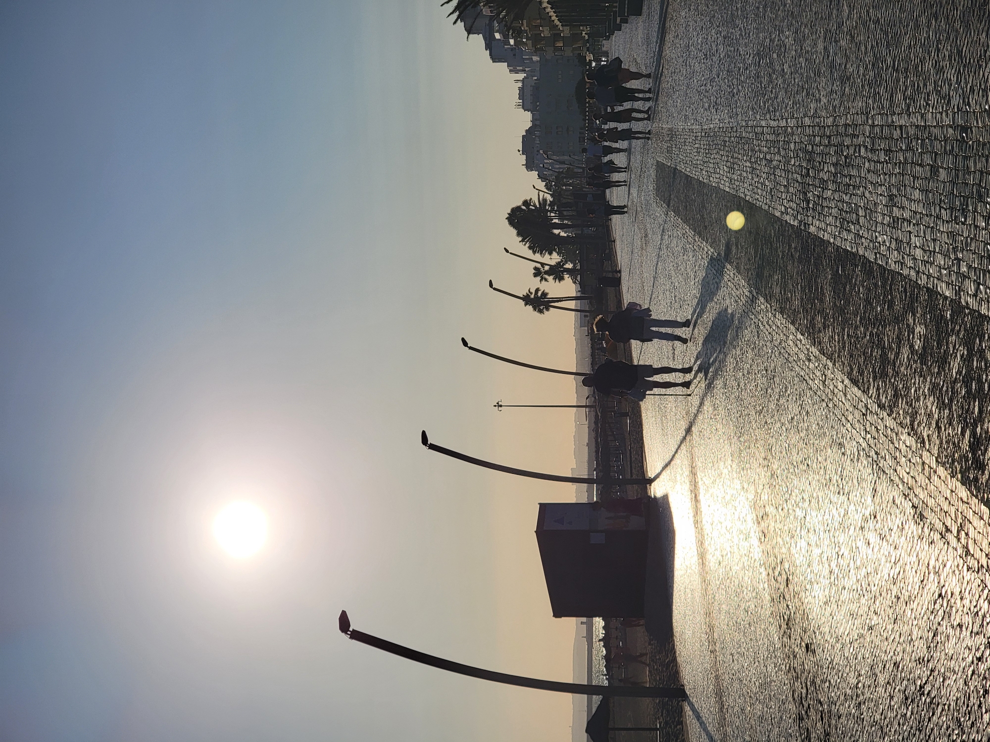 The image depicts a sunny day at a cobblestone promenade in Quarteira, Portugal. The sun is high in the sky, casting long shadows and creating a bright glare on the walkway. Several people are walking along the promenade, some singly and others in small groups. There are distinctive curved lampposts lining the promenade, and a dark rectangular structure, possibly a kiosk or public facility, is visible on the left side of the image. Beyond the promenade, a beach with umbrellas and people can be seen, leading to the ocean. In the background, buildings and palm trees suggest a coastal urban environment. The scene conveys a sense of leisure and activity on a pleasant day.