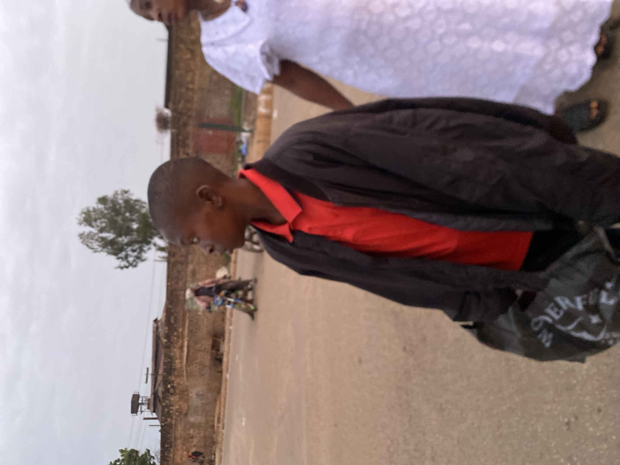 An eye-level, medium shot shows two people walking on a paved road in what appears to be Bukuru, Nigeria. The primary subject is a man wearing a red collared shirt covered by a black jacket; he is carrying a dark-colored bag with some white lettering. The second person, partly visible in the upper frame, is wearing a white lace dress. The background includes buildings of local architecture, a tree, and a small motorcycle traveling on the road. The sky is overcast.