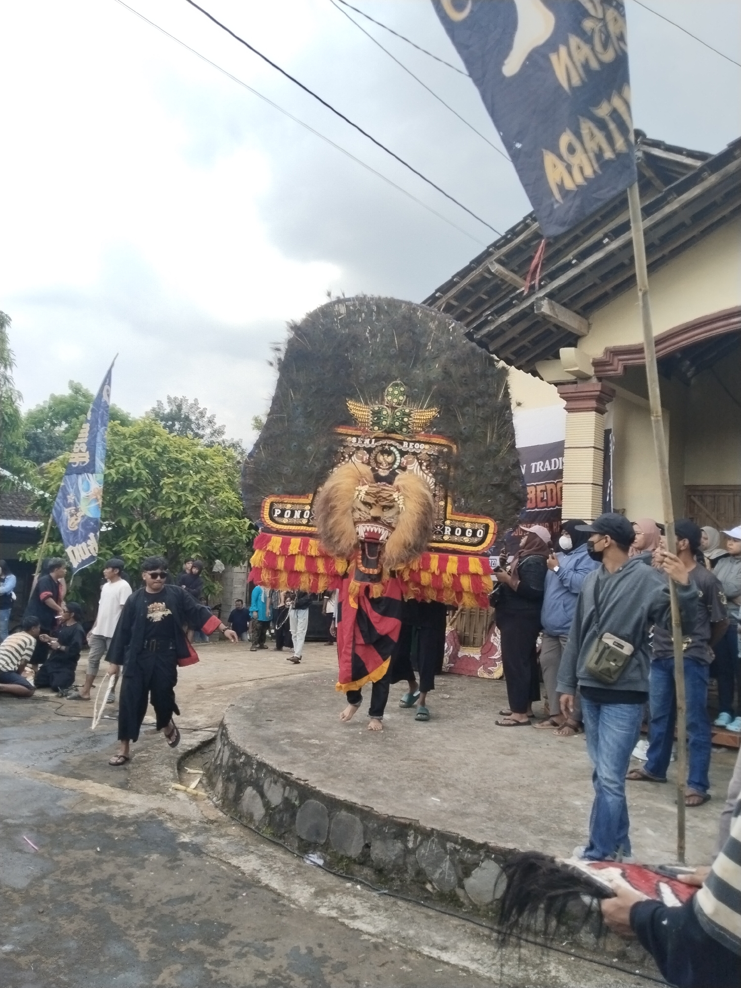 Captured in Pendem, Indonesia, the image shows a traditional cultural performance taking place outdoors, with a crowd gathered around. The central focus is a large, elaborate headdress resembling a lion's head with a mane of feathers and colorful embellishments. Performers hold the headdress aloft, moving along a stone platform. Other performers and spectators stand nearby, some wearing casual clothing and face masks. Banners and flags are present, adding to the festive atmosphere. The surrounding environment includes a house with a traditional roof, trees, and a paved area.