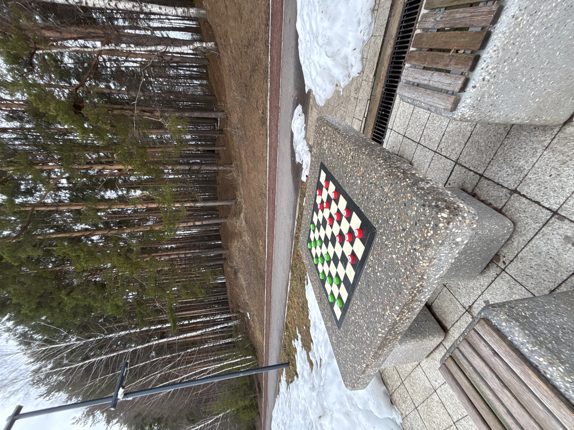 The image shows a checkers table in a park in Chaikovsky, Russia. The table is made of speckled concrete, with a black and white checkerboard inlay. Red and green checker pieces are arranged on the board. Two wooden benches flank the table. The ground around the table is paved with light-colored tiles and patches of snow. In the background is a forest and a paved path. The trees are bare-branched, indicating it is likely late fall or winter.