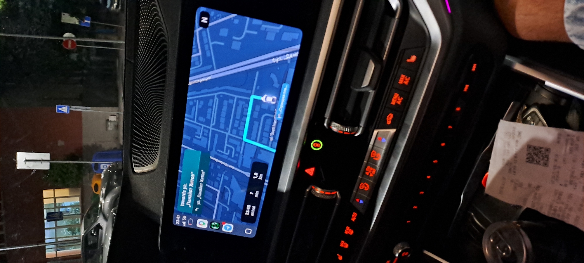 The image is a close-up interior shot of a car at night, focused on the dashboard and navigation screen. The navigation screen displays a map of Burgas, Bulgaria, with a route highlighted. The destination is indicated as "ул. Панайот Хитов" (Panayot Hitov Street). The time on the navigation system is 23:48, with an estimated arrival in 7 minutes, covering a distance of 1.8 km.
The car's climate control panel is visible below the navigation screen, with illuminated buttons indicating various functions like heated seats and AC settings. A hazard light button is prominently displayed at the top of the panel. To the left of the hazard button, a green circular light is illuminated, possibly indicating a system status.
Outside the car, through the windshield, a street scene at night is visible. Streetlights and building lights illuminate the area. A dark-colored car is parked on the left, and a blue trash bin is visible next to it. Traffic signs, including a pedestrian crossing sign and a circular red sign, are present on the street.
In the foreground, inside the car, a crumpled receipt is visible in the center console, along with car keys and a soda can. The overall impression is that of a car driving at night in an urban environment, with the navigation system guiding the journey through Burgas.