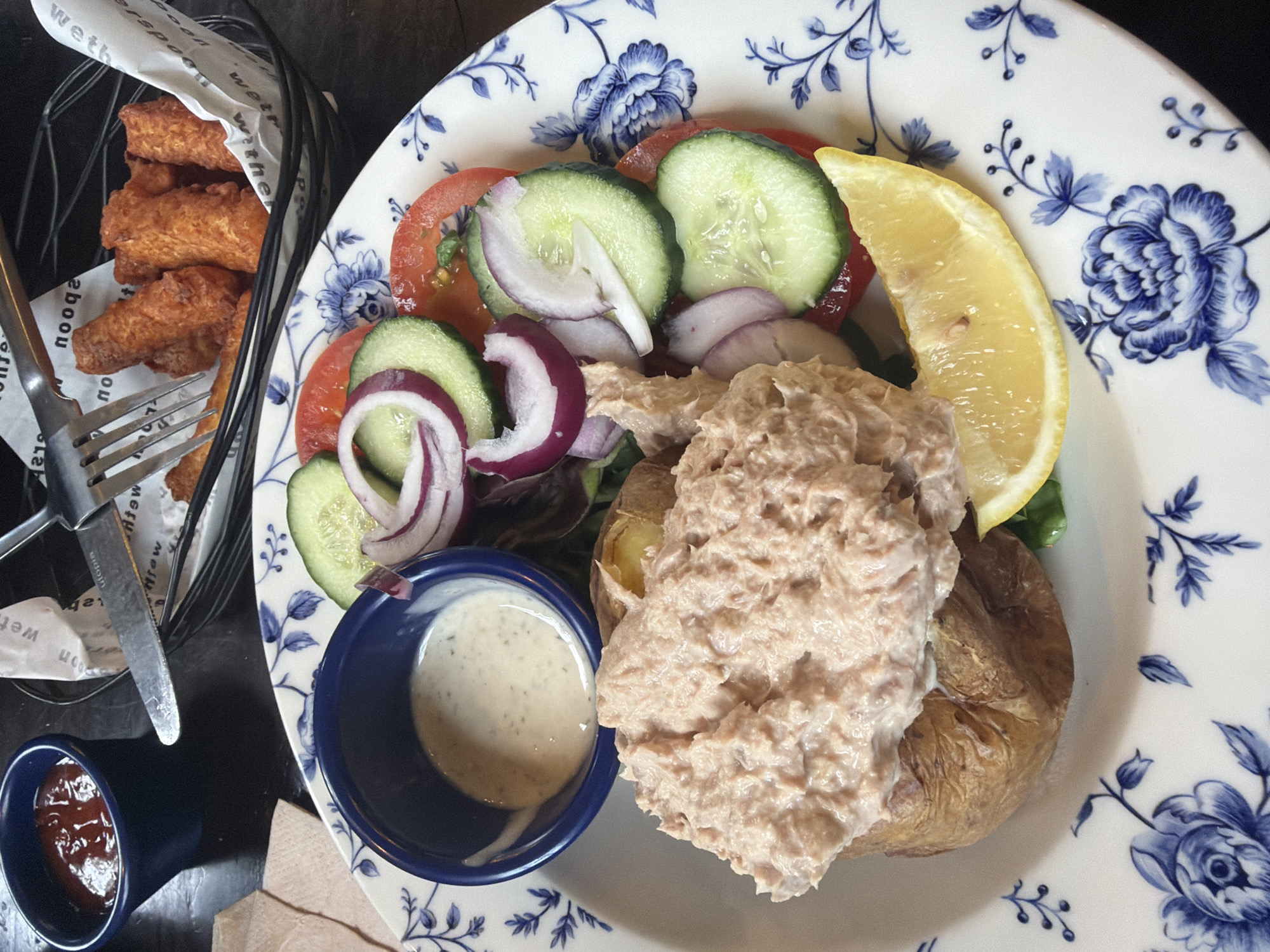 A close-up, eye-level shot displays a meal on a decorative white plate with blue floral patterns. The meal consists of a baked potato topped with a generous portion of tuna mayonnaise, a side salad with tomatoes, cucumbers, and red onion, a wedge of lemon, and a small blue bowl of salad dressing. In the background, another blue bowl holds ketchup, and a black basket contains several pieces of fried fish sticks, with a fork and knife resting on white paper above them. The setting appears to be a restaurant or pub in Epsom and Ewell, United Kingdom, suggested by the style of the food and presentation. The lighting is natural and ambient, highlighting the textures and colors of the food.