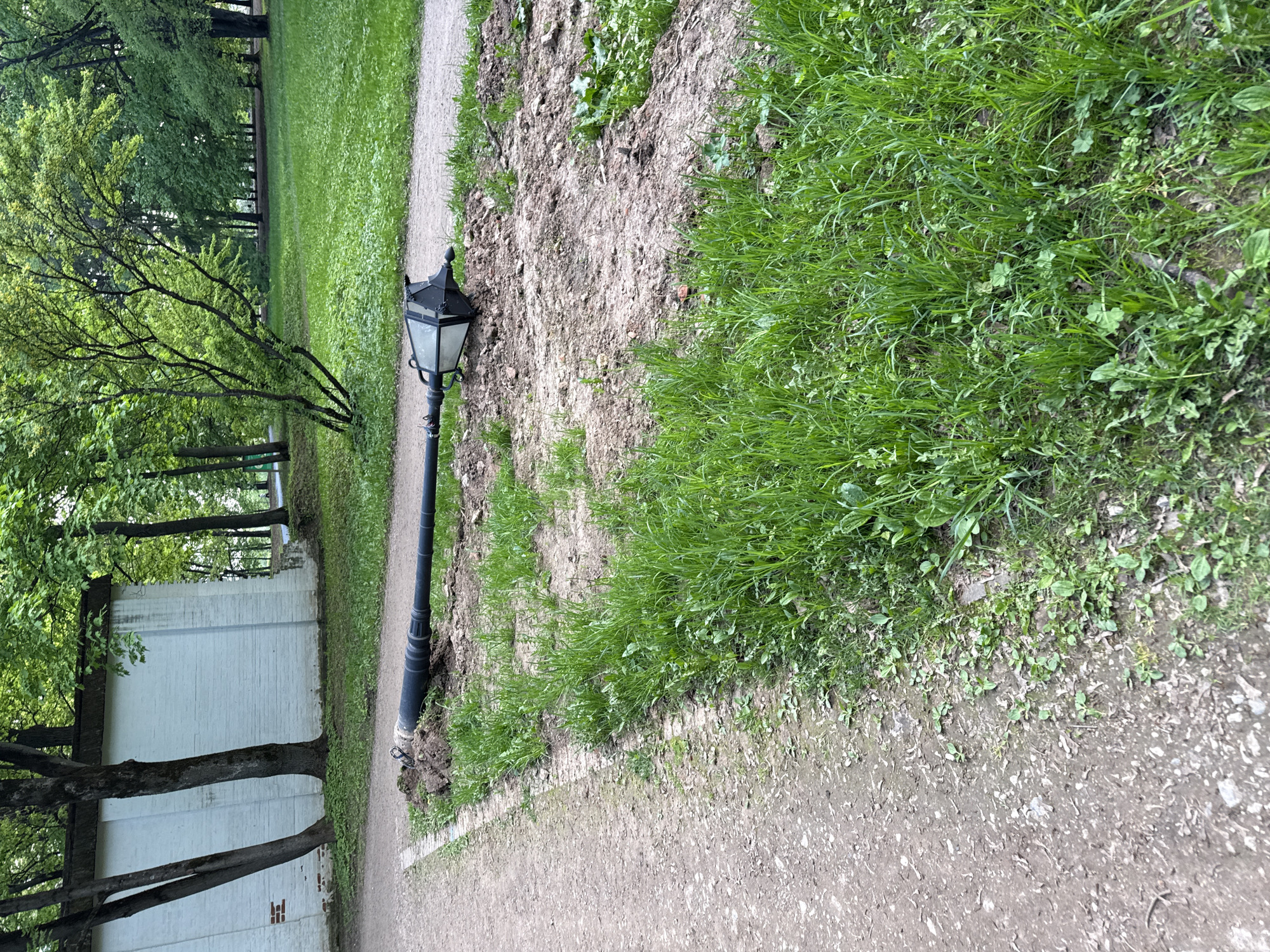 The image shows a park in Moscow, Russia. A dirt path runs along the bottom edge of the frame. A vintage black lamppost stands beside the path. There is an unkempt strip of grass and soil next to the path. A white brick wall runs parallel to the path on the left side of the frame, with trees growing nearby. Beyond the wall and to the top left of the frame are more trees and green grass.