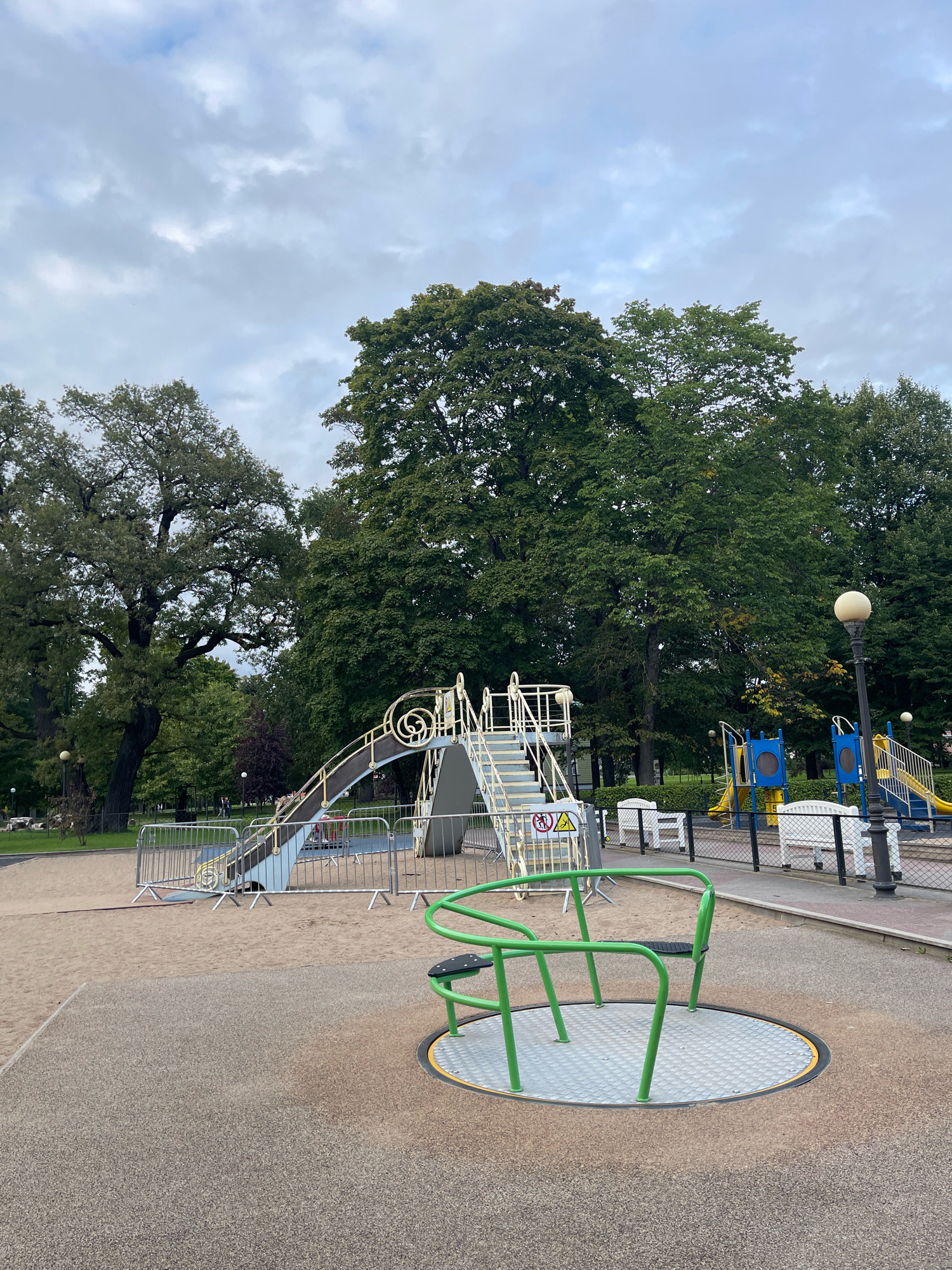 The image shows a playground in Tallinn, Estonia. The playground is equipped with a slide, a merry-go-round, and a swing set. The ground is covered with sand and rubber mulch. There are several trees in the background, and the sky is cloudy. The image is taken from a low angle, and the playground is in focus.