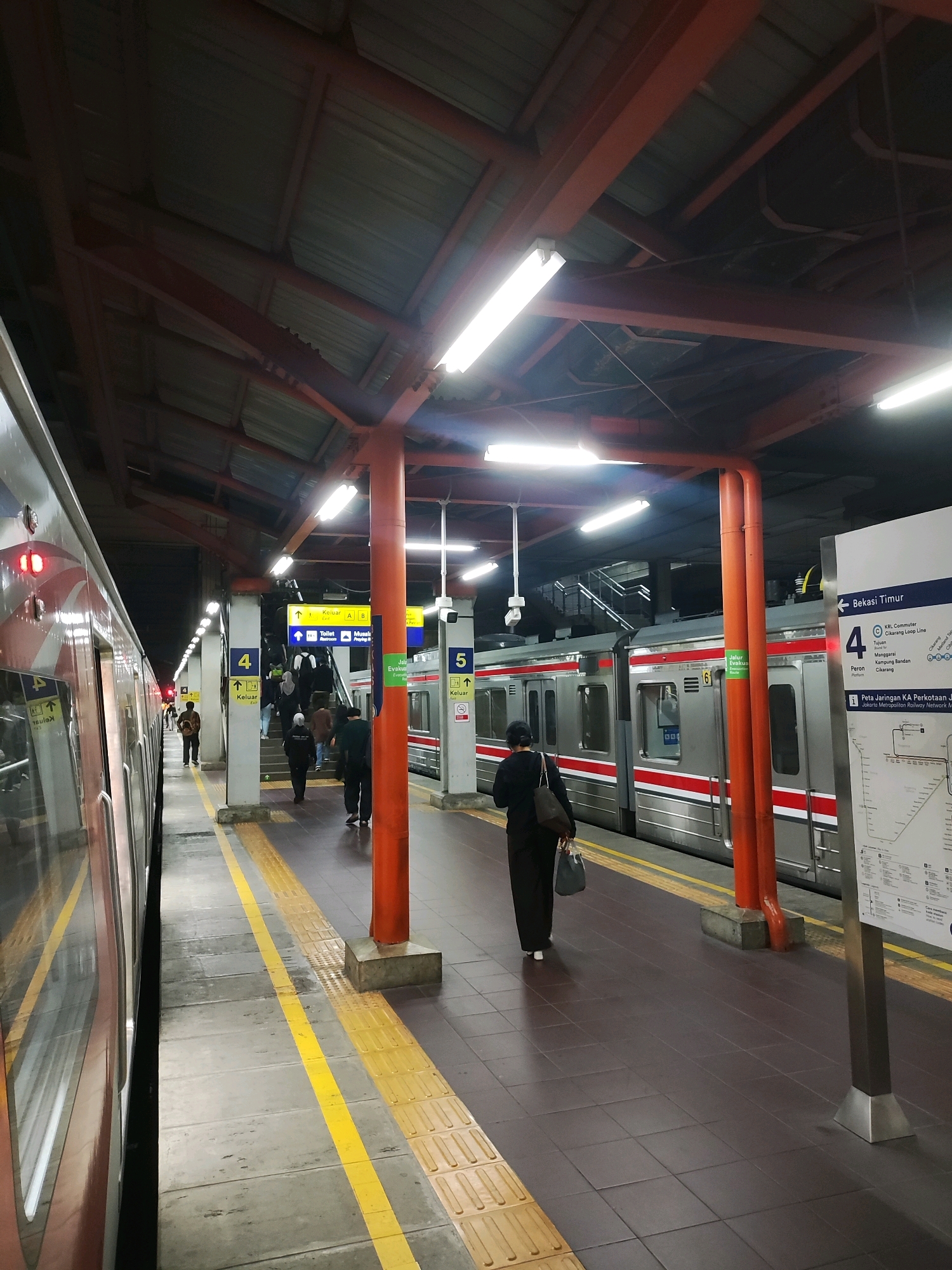 The image captures a platform at a train station in Bekasi, Indonesia. A train with red and silver cars is parked on the left side of the frame, its reflection visible on the polished surface of the train. On the platform, several people are walking in various directions. One woman in black is walking away from the camera, carrying two bags. Another person in a black jacket is walking towards the camera. In the background, a staircase leads upwards. Overhead, a corrugated metal roof supported by orange beams covers the platform. Several fluorescent lights illuminate the scene. Yellow tactile paving runs along the edge of the platform, and yellow lines mark the safe waiting area. Signs with Indonesian text and arrows are posted, indicating platform numbers and directions. A large information board with a map of the Jakarta Metropolitan Railway Network is visible on the right side of the image. The overall atmosphere suggests a busy public transport hub.