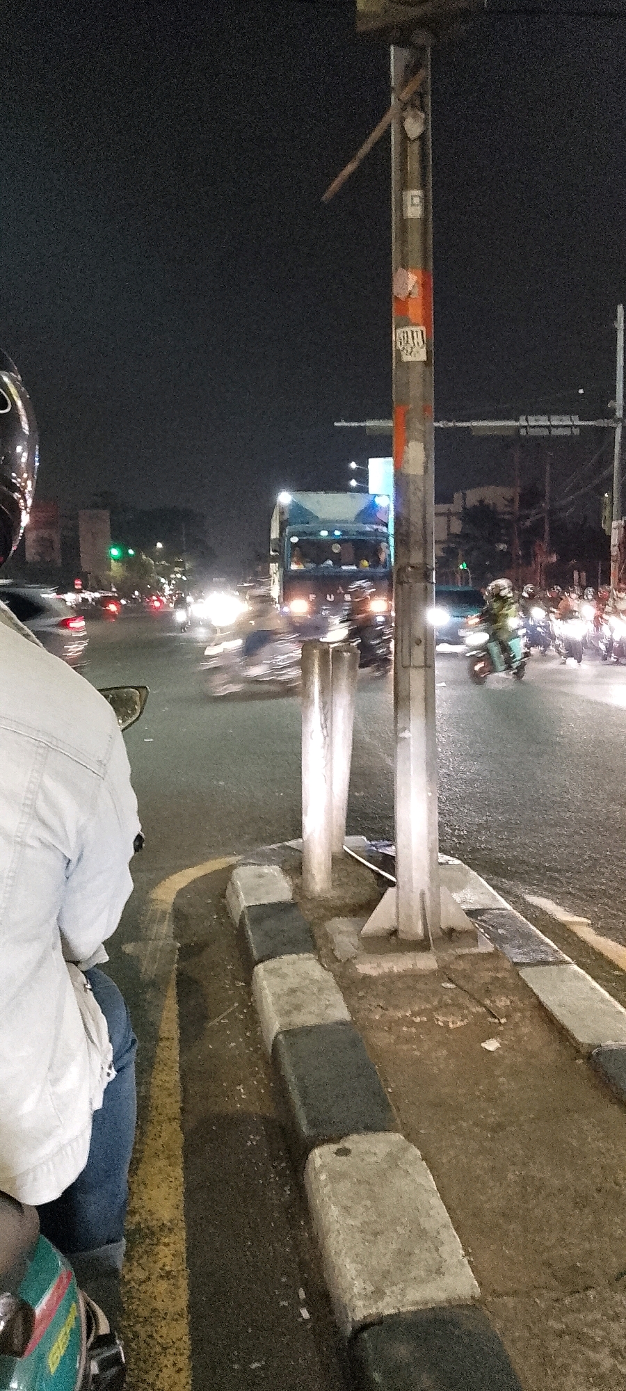 The image captures a night scene at an intersection in Depok, Indonesia. The perspective is from someone seated on a motorcycle, looking towards the traffic. In the foreground, the shoulder of a person wearing a light blue denim jacket and blue jeans is visible, along with a portion of a motorcycle's front fender. The road is wet and reflects the lights of vehicles.
The intersection is busy with vehicular traffic, including a large blue truck with illuminated headlights and a group of motorcycles with their lights on. Some vehicles are blurred due to motion. Traffic lights are visible in the background, with one showing a green signal. A utility pole, with stickers and some orange markings, stands prominently in the mid-ground. The environment is characterized by darkness, illuminated by streetlights and vehicle headlights, indicating a typical urban night setting in Depok.