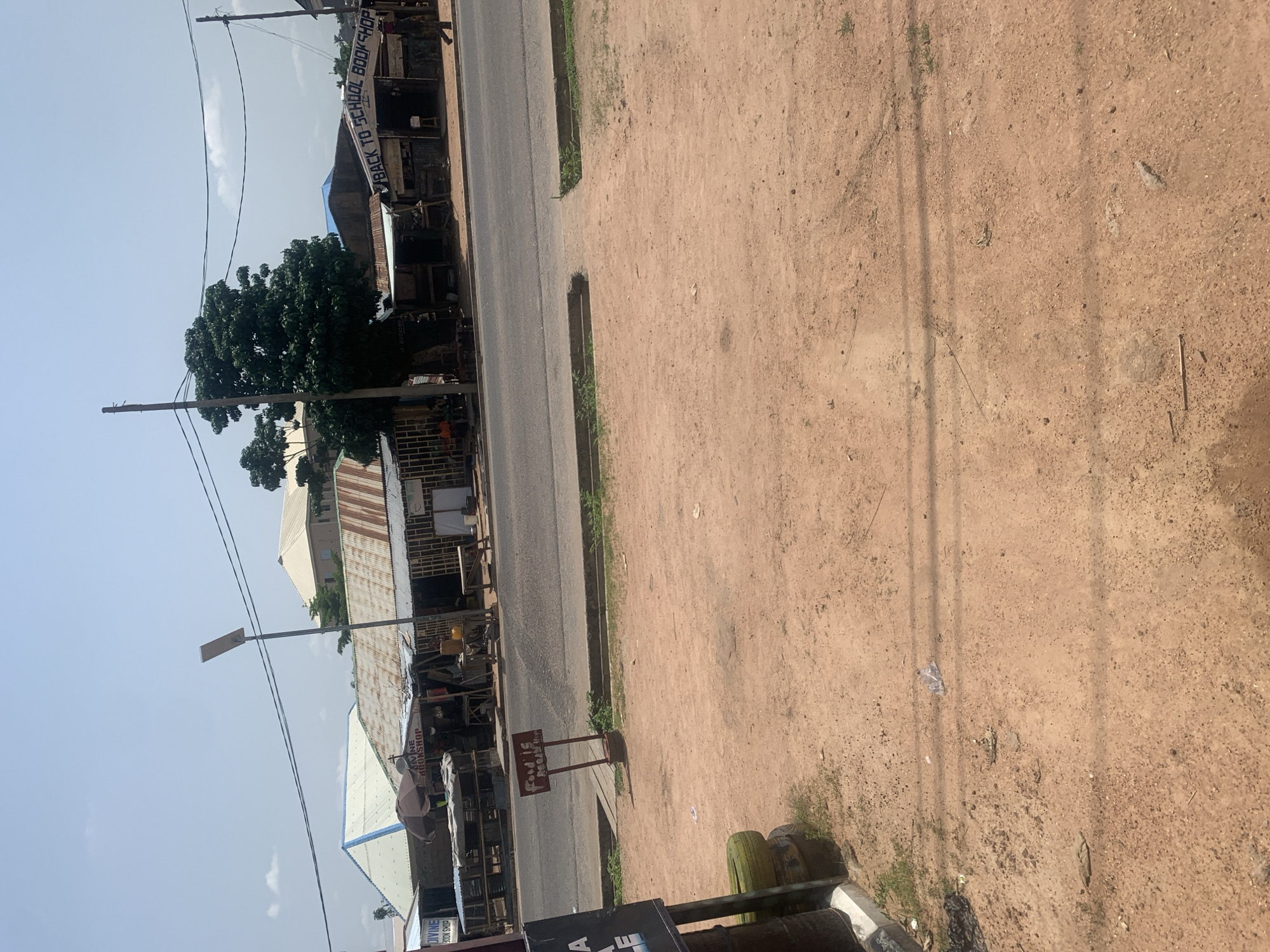 This image depicts a street scene in Jalingo, Nigeria. The primary subjects include a paved road, a dusty dirt shoulder, and several small shops or market stalls lining the roadside. A prominent feature is a tall tree with lush green foliage situated between the road and the shops. Power lines crisscross the sky, and a street lamp is visible.
There are no discernible people actively engaged in an event in the frame. The activity appears to be a still moment in a commercial area, possibly during business hours, though the lack of people makes it difficult to ascertain.
Notable details include the dry, sandy texture of the ground on the shoulder, suggesting a warm and possibly dry climate. The buildings have corrugated metal roofs, a common architectural style in many regions. The sky is a pale blue with a few wispy clouds, indicating daytime, likely mid-morning or afternoon, with clear weather.
Visible text includes "BACK TO SCHOOL BOOK SHOP" on a banner hanging over one of the shops. Another sign, a small wooden one on a stand, has the text "God is Peace" visible. Additionally, part of the text "DIVINE BOOKSHOP" is visible on a sign. The overall impression is a slice of everyday life in a bustling commercial area of Jalingo.