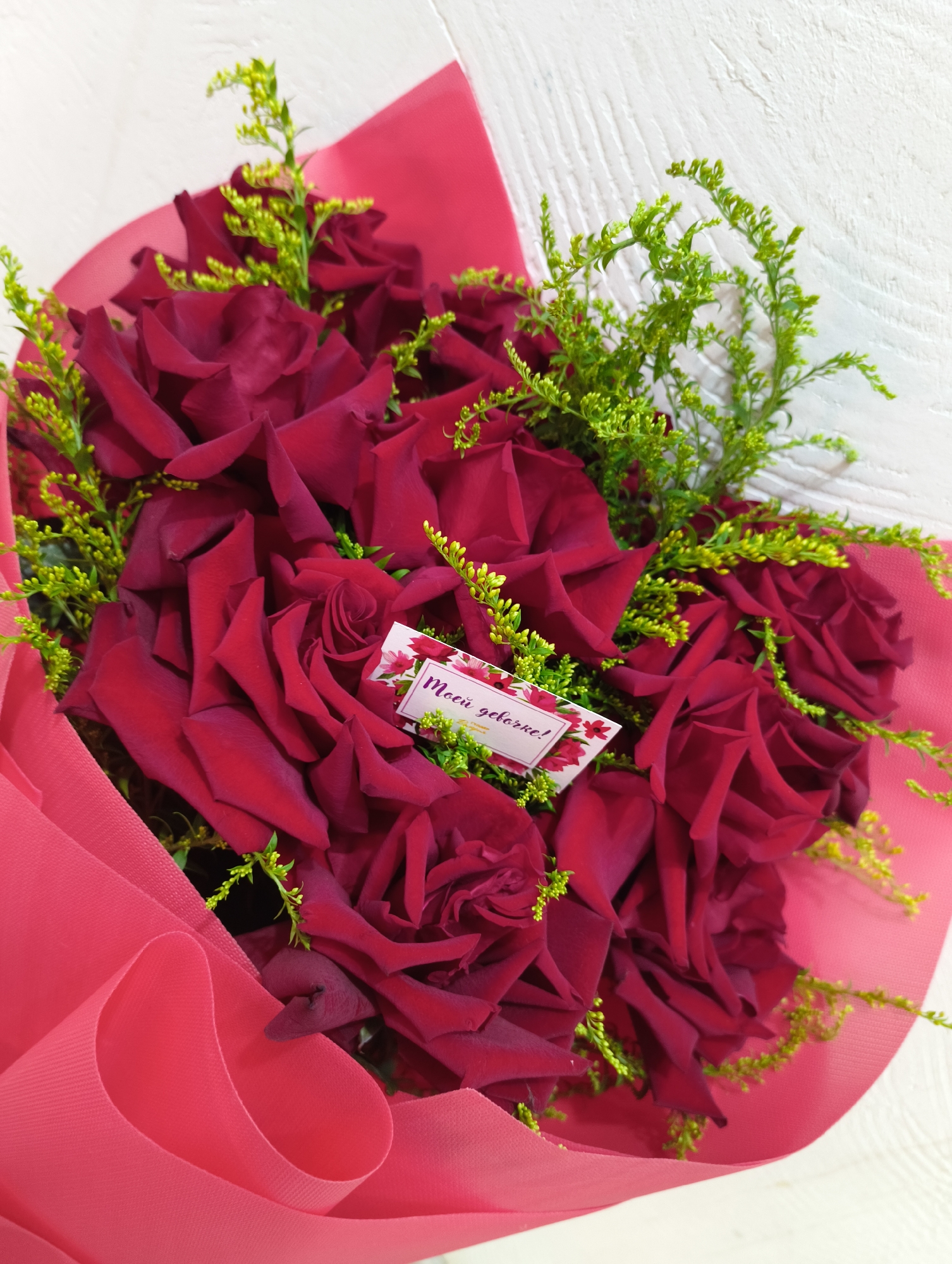 A bouquet of deep red roses is displayed against a white, textured background. The roses are lush and velvety, with some subtle variations in shade creating depth. They are interspersed with delicate, bright green filler flowers and stems that add a pop of contrasting color and texture. The bouquet is partially wrapped in a vibrant pink paper, creating a soft, layered effect. A small, rectangular card with a floral border is visible, featuring text in a dark color that appears to be Cyrillic. The card is placed amongst the rose petals. The overall impression is one of romantic elegance and thoughtful presentation.