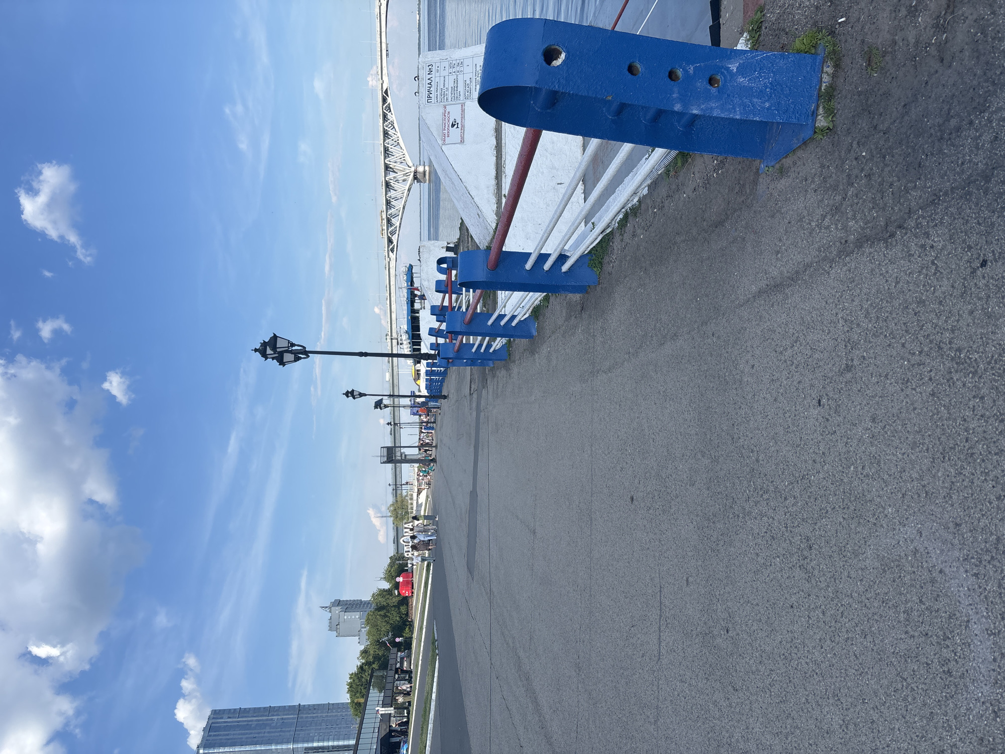 The image captures a waterfront promenade in Saratov, Russia, featuring a paved walkway alongside a body of water. A blue and white railing with red accents delineates the edge of the walkway. In the background, a large bridge spans the water, and several buildings, including a tall, modern skyscraper, are visible on the skyline. Several people are scattered along the promenade, some walking, others standing. The sky is blue with scattered clouds, indicating fair weather. A street lamp is also visible along the walkway. A white structure with Russian text, possibly a sign or informational board, is positioned on the embankment. The overall scene depicts a public space during daylight hours.