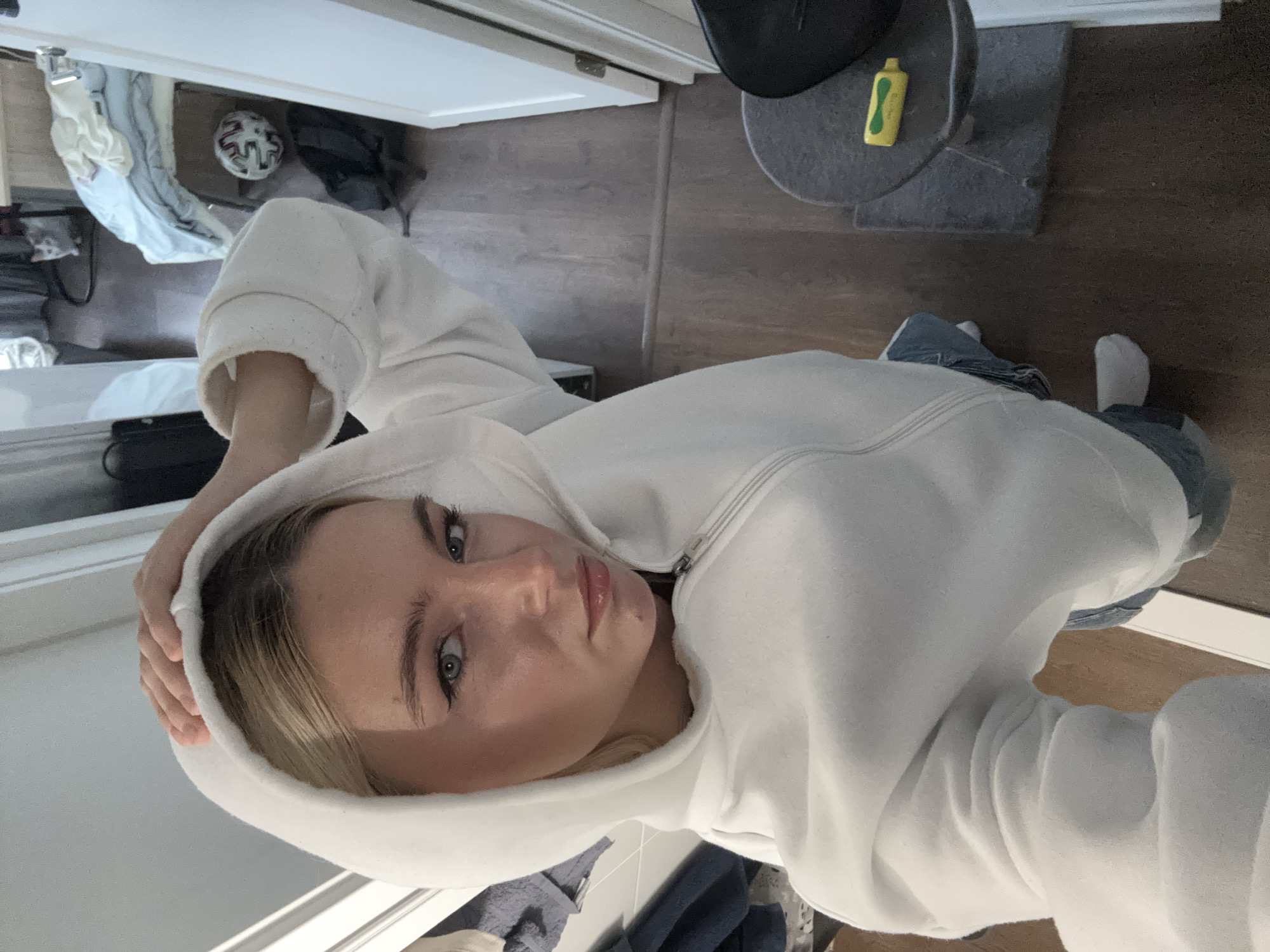 A selfie of a young woman with light skin and blonde hair wearing a white zip-up hoodie. The background includes a room with a door, a cat tower with a black hat and yellow bottle on it, and some clothing items scattered on the floor. The location is not discernible.
