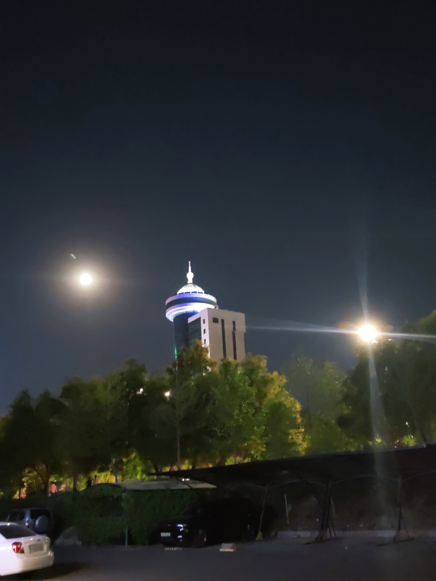 The image is a night-time photograph taken in Tashkent, Uzbekistan, featuring the Tashkent TV Tower. The moon is visible in the dark sky, casting a soft glow. The Tashkent TV Tower, illuminated with blue and white lights, stands prominently in the background. In the foreground, trees are visible, with some parking areas and a few cars. The overall scene depicts a night view of a cityscape with a notable landmark.