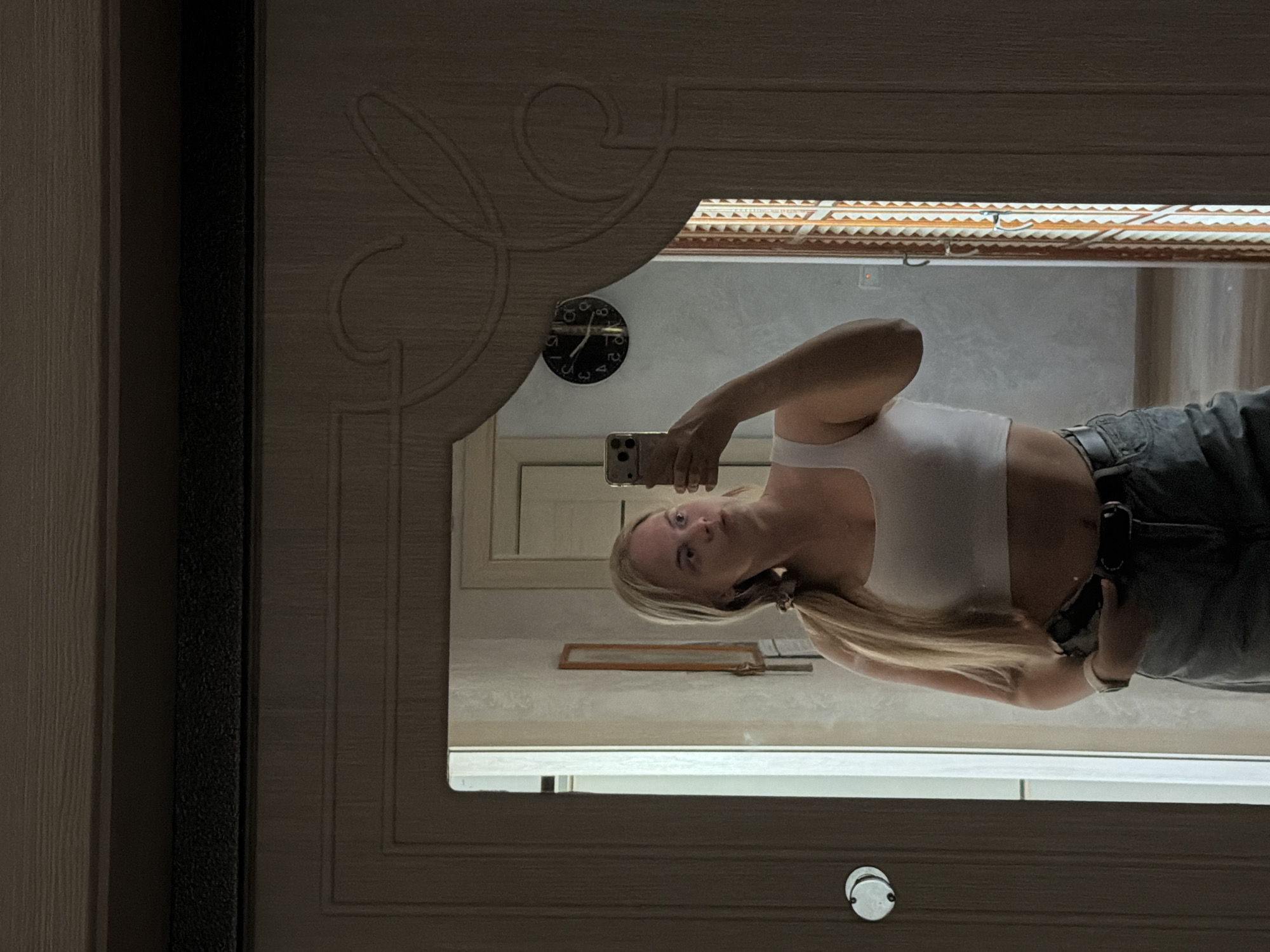 A medium shot shows a mirror in a door frame reflecting a blonde woman taking a selfie with a smartphone. The woman is wearing a white tank top and jeans. The background appears to be a room inside a building, with an interior design characteristic of residences. In the upper-left corner of the reflection, a clock indicates the time to be approximately 8:20. The scene likely takes place inside a residential building in the city of Chaikovsky, Russia, based on prior knowledge.
