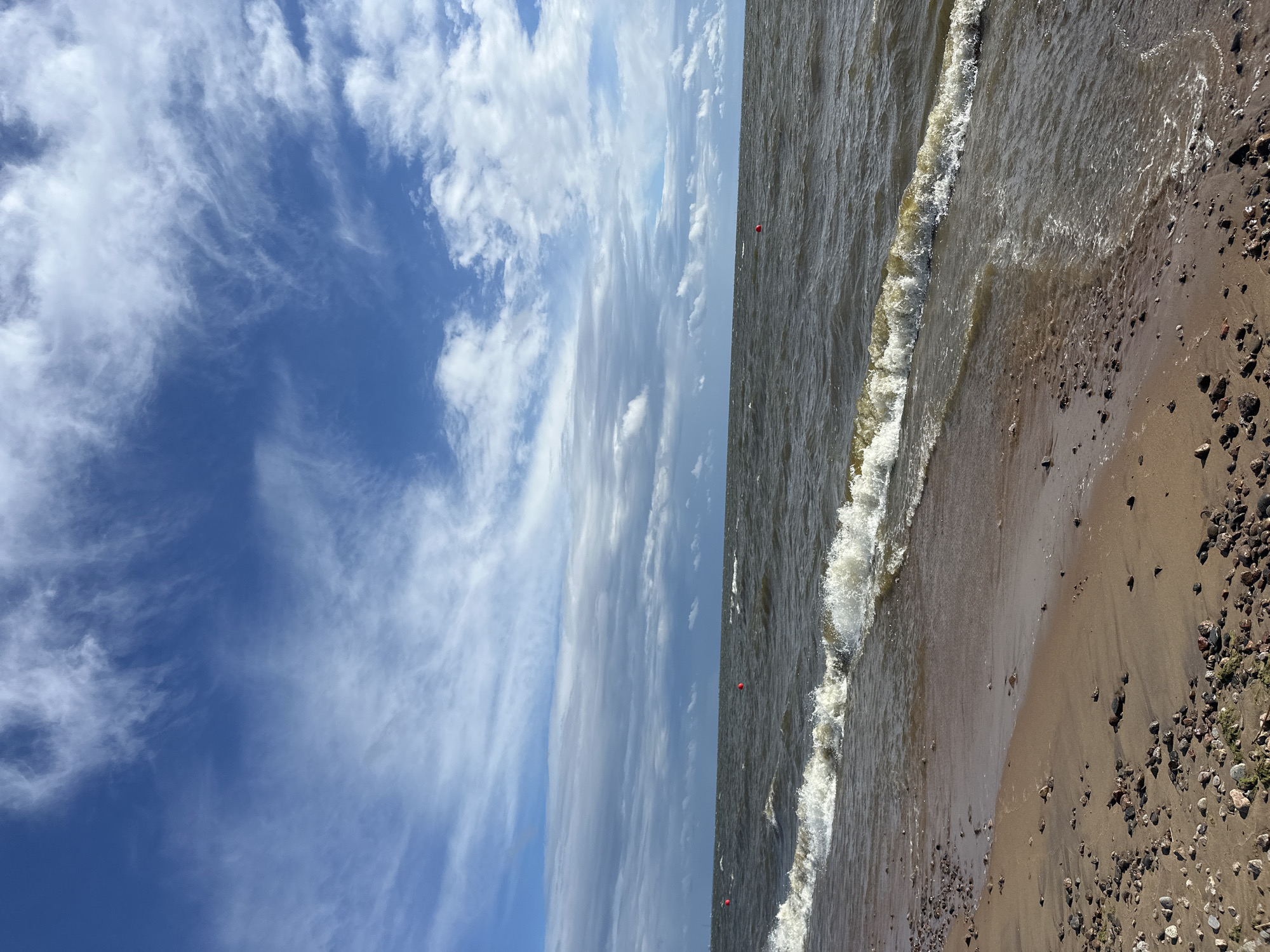 This is a photograph of a beach scene in Zelenogorsk, Russia, on a partly cloudy day. The image captures the sandy shore, waves lapping at the edge, and the sea stretching to the horizon under a sky with wispy clouds.
The foreground features a sandy beach littered with small pebbles and some dried seaweed. A gentle wave is breaking on the shore, with white foam and clear water visible. The sea is choppy with moderate waves, appearing a dark, murky green-brown. In the distance, three red buoys are visible in the water.
The sky is a vibrant blue, populated by scattered white, wispy clouds that are elongated and streaky, suggesting wind. A darker band of clouds runs along the horizon, creating a contrast with the brighter sky above.
There are no people or animals depicted in the image. The scene is a natural landscape photograph focused on the elements of the beach, sea, and sky.
