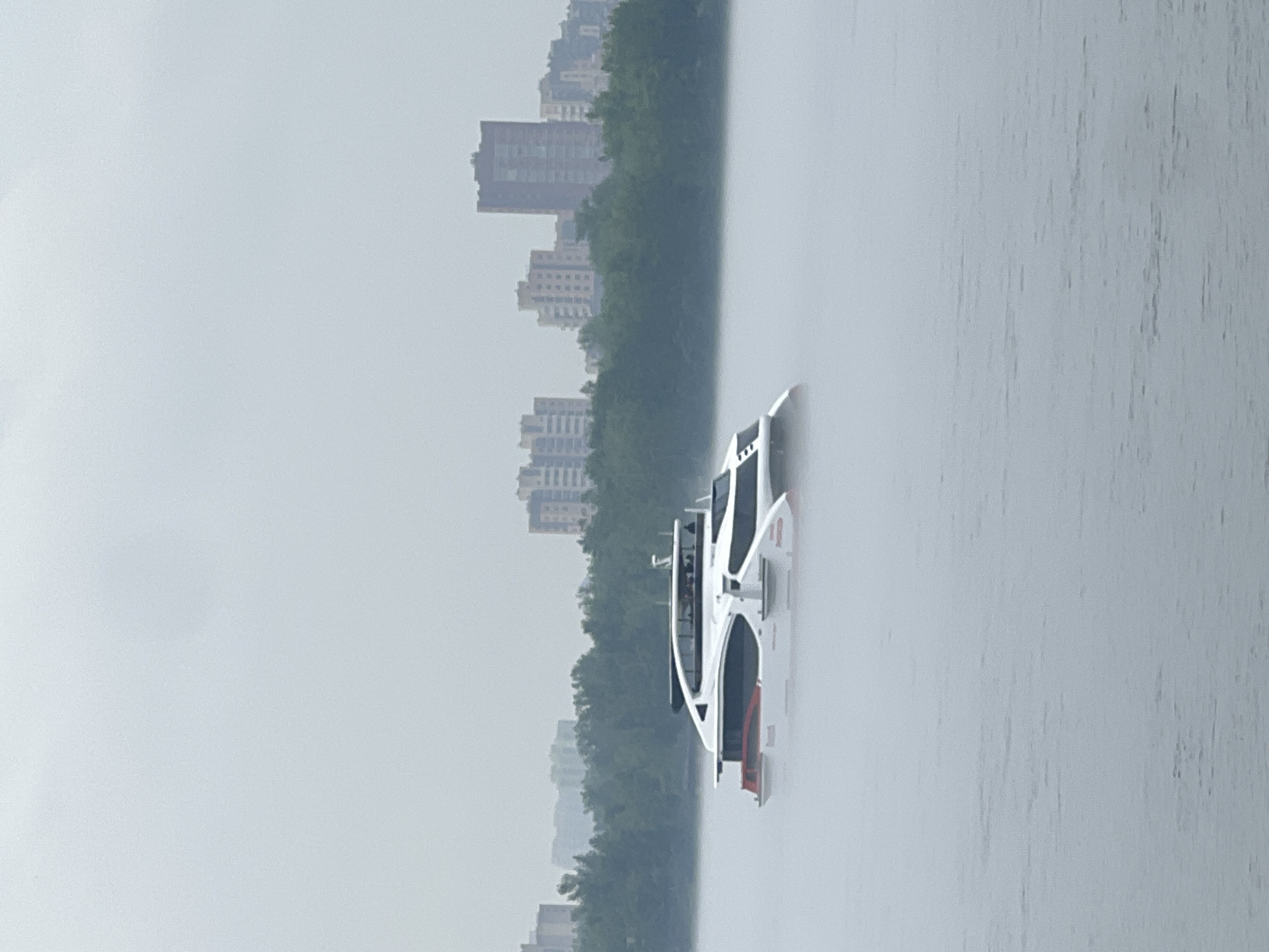 A modern, white passenger catamaran with red accents is navigating a foggy river in Krasnoyarsk, Russia. The boat, which appears to be a hydrofoil or high-speed ferry, has a sleek design with multiple levels and large windows. People are visible inside the cabin. The river is shrouded in dense fog, obscuring the water's surface and much of the surrounding environment. In the background, a line of lush green trees forms a dense forest along the riverbank. Behind the trees, a hazy silhouette of apartment buildings and urban structures can be seen, suggesting the presence of a city. The sky is overcast and uniformly gray, contributing to the overall muted and diffused lighting of the scene. The image captures a moment of low visibility, where the boat emerges from the mist against the backdrop of the city and natural landscape.