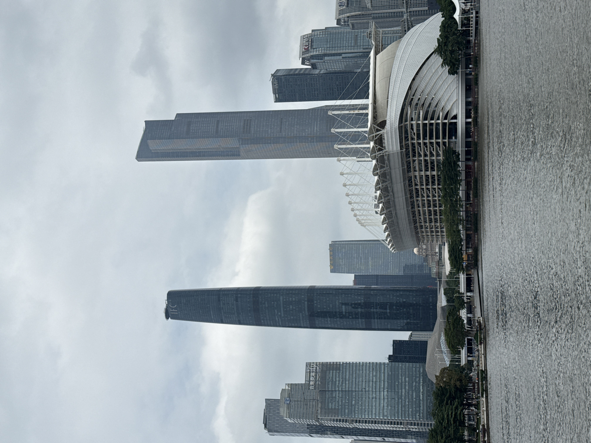 The image captures a cityscape in Yuexiu District, China. Several skyscrapers rise in the background, including one with the label "IFC" visible at its top. Near the water's edge is the Guangzhou Opera House, characterized by its modern architectural design with a curving, angular roof and white support structures. The foreground features a body of water, likely a river, with small ripples visible on its surface. Trees and possibly a promenade line the edge of the water. The sky is overcast, suggesting a cloudy day.