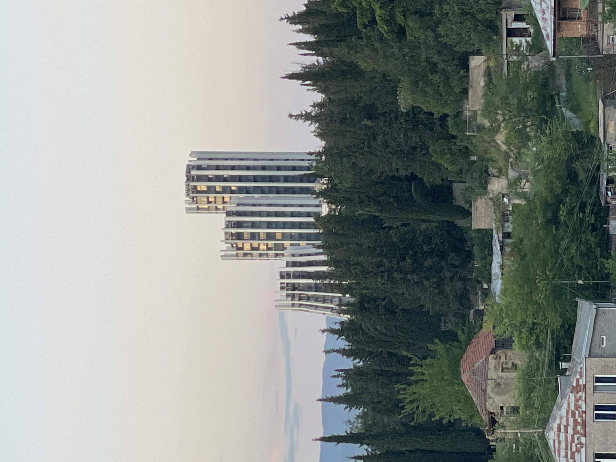 The image shows a cityscape in Tbilisi, Georgia. In the background, there is a modern white building with several stories, possibly a residential or commercial complex. To the right of the building, there is a dense forest. Closer to the foreground, there are older, run-down buildings mixed in with trees and vegetation. The sky is overcast, and the image appears to have been taken at dusk or dawn. No people are visible in the image.