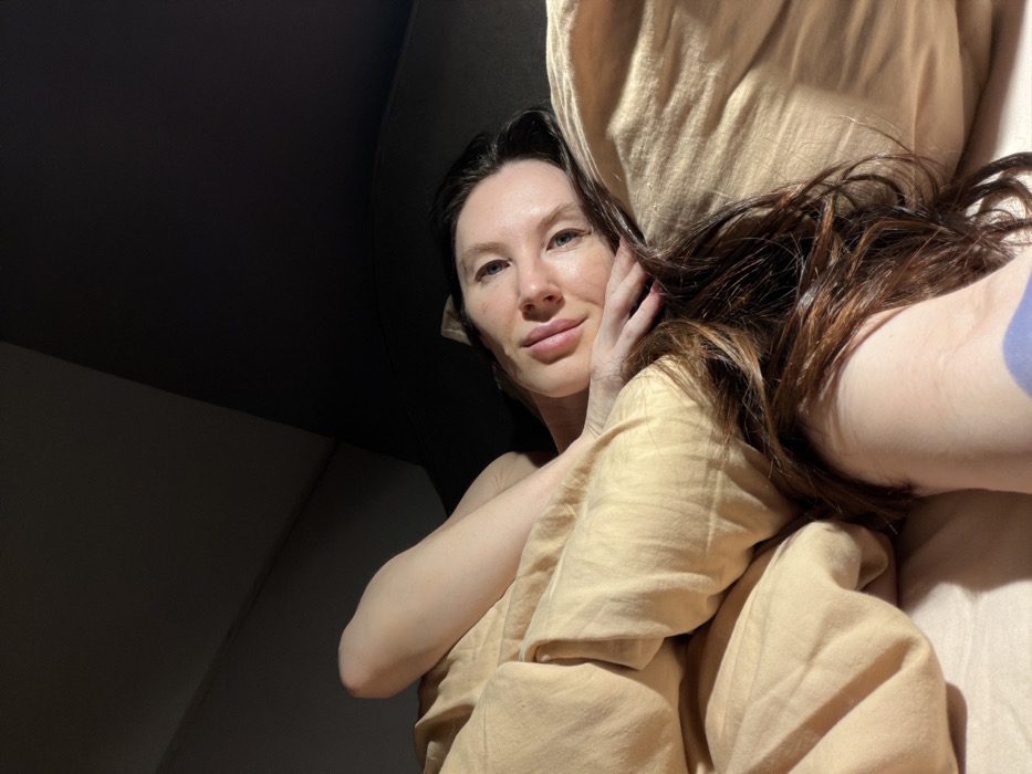 A woman with dark hair and fair skin poses in bed. She is partially covered by a light yellow blanket and pillow. Her head rests on her hand, and she smiles softly at the camera. The background is mostly dark, with the corner of the room visible. The image is taken from a low angle, looking up at the woman. Her hair appears damp, and the scene is illuminated by natural light.