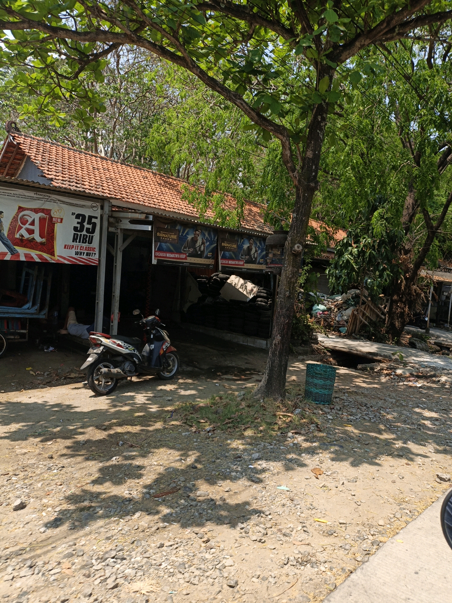 In Ujungbatu, Indonesia, a roadside scene unfolds under a canopy of green trees. A red-tiled roof structure, likely a shop or food stall, is prominent. A white banner with red lettering, partially obscured, reads "35 RİBU KEEP IT CLASSIC. WHY NOT?". A motorcycle is parked on a gravelly ground in front of the structure. To the left, a person lies down, partially covered, near what appears to be a colorful truck or cart. The background reveals more greenery and some piles of discarded materials. Two posters are affixed to the building, one featuring a man labeled "SIGNATURE" and the text "RASAKAN NIKMATNYA HIDUP". A blue-green woven container is visible on the ground near a tree trunk, casting shadows on the textured ground. The environment suggests a warm, possibly humid, tropical climate.