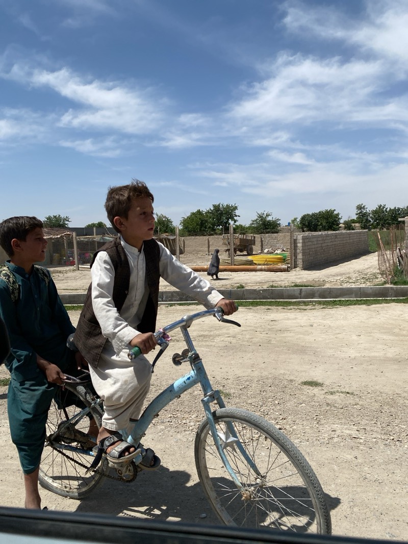 Two boys are on a bicycle on a dirt road in Kandahar, Afghanistan. The boy in front is riding the bicycle and wears a white salwar kameez with a brown vest and sandals. The boy behind him is sitting on the back rack of the bicycle and wears a dark green salwar kameez. A woman in a black burqa walks along the dirt road in the background. Yellow pipes are stacked near a low brick wall in the background. The sky is light blue with white clouds.