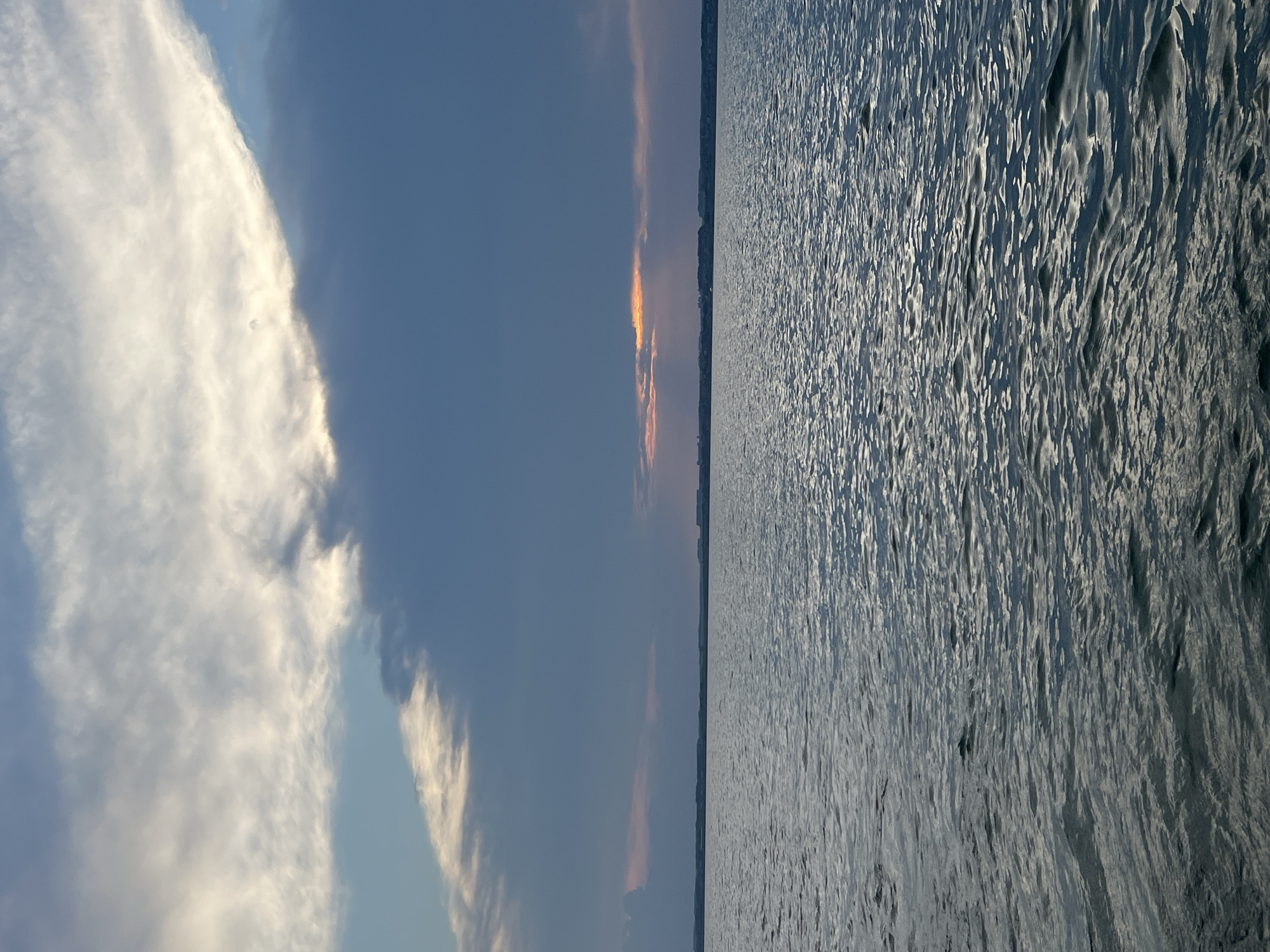 The image shows a body of water with waves reflecting the sky. The sky is filled with clouds, some of which are illuminated by sunlight breaking through. In the distance, a faint line of land with some structures is visible along the horizon. The scene appears to be a natural landscape, possibly a lake or the sea, under a dramatic sky. There are no people, animals, or man-made objects in the immediate foreground or midground. The overall mood is serene and natural. There is no information in the image to suggest the location is in the city of Novopichugovo, Russia.