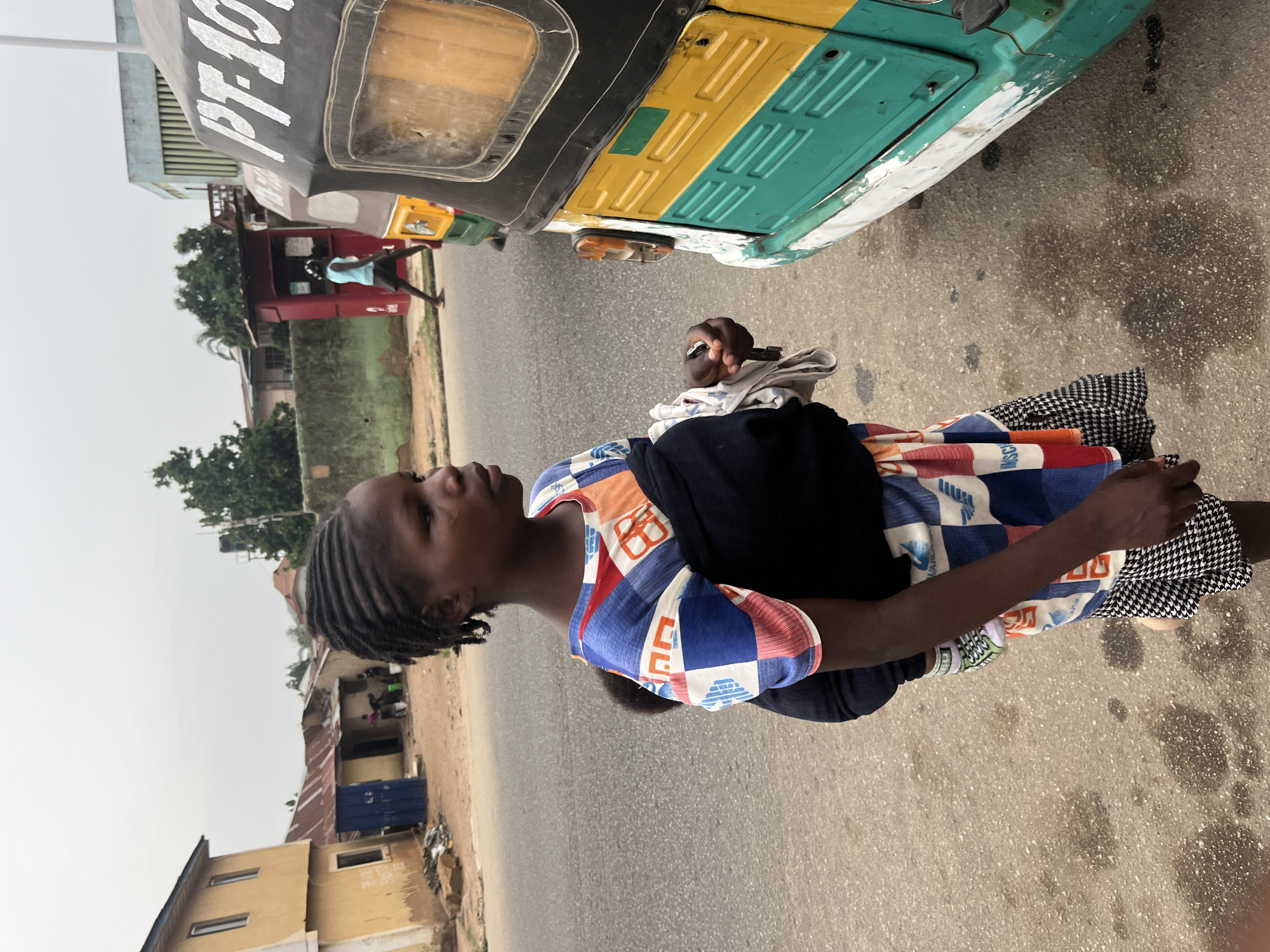 A young African woman with braided hair stands on the roadside in Jos, Nigeria, near a yellow and green taxi with registration number "PF-1076". She wears a colorful patterned dress with a dark top, and a black and white patterned skirt. She holds a metal object in her right hand and a white cloth in her left. The road is gray and shows signs of wear. Buildings and trees line the street in the background, and a man in a blue shirt walks by a red kiosk. The sky is overcast.