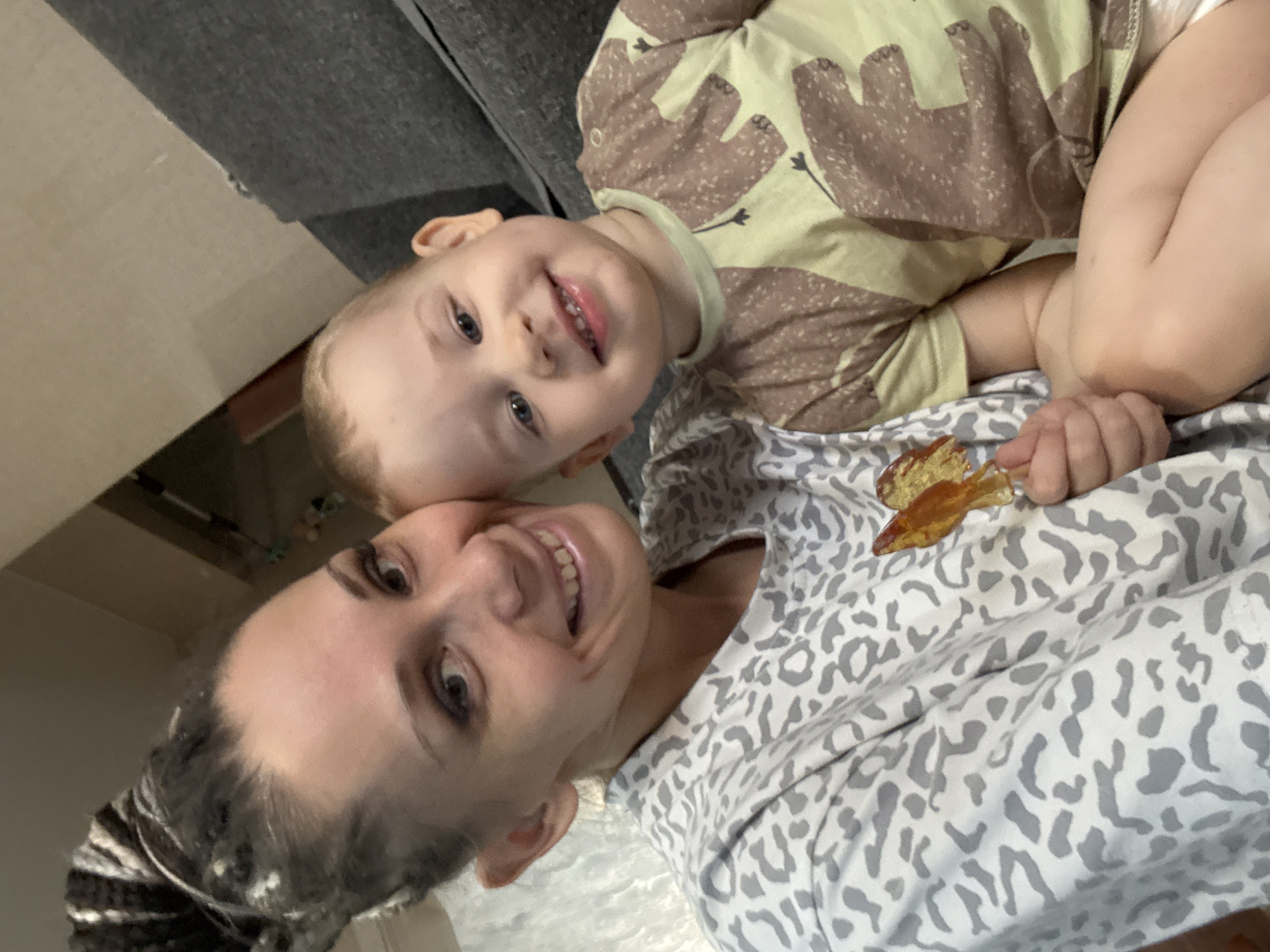The image shows a selfie of a woman and a young child, presumably in a domestic setting. The woman is at the bottom of the frame, lying down with her head tilted towards the camera, smiling. She has her hair pulled back. The child is lying on top of her, also smiling, with a light-colored shirt featuring a brown animal pattern. The background includes a portion of a gray couch and a wall. The environment suggests an informal, indoor setting. The location is Unknown, Unknown.