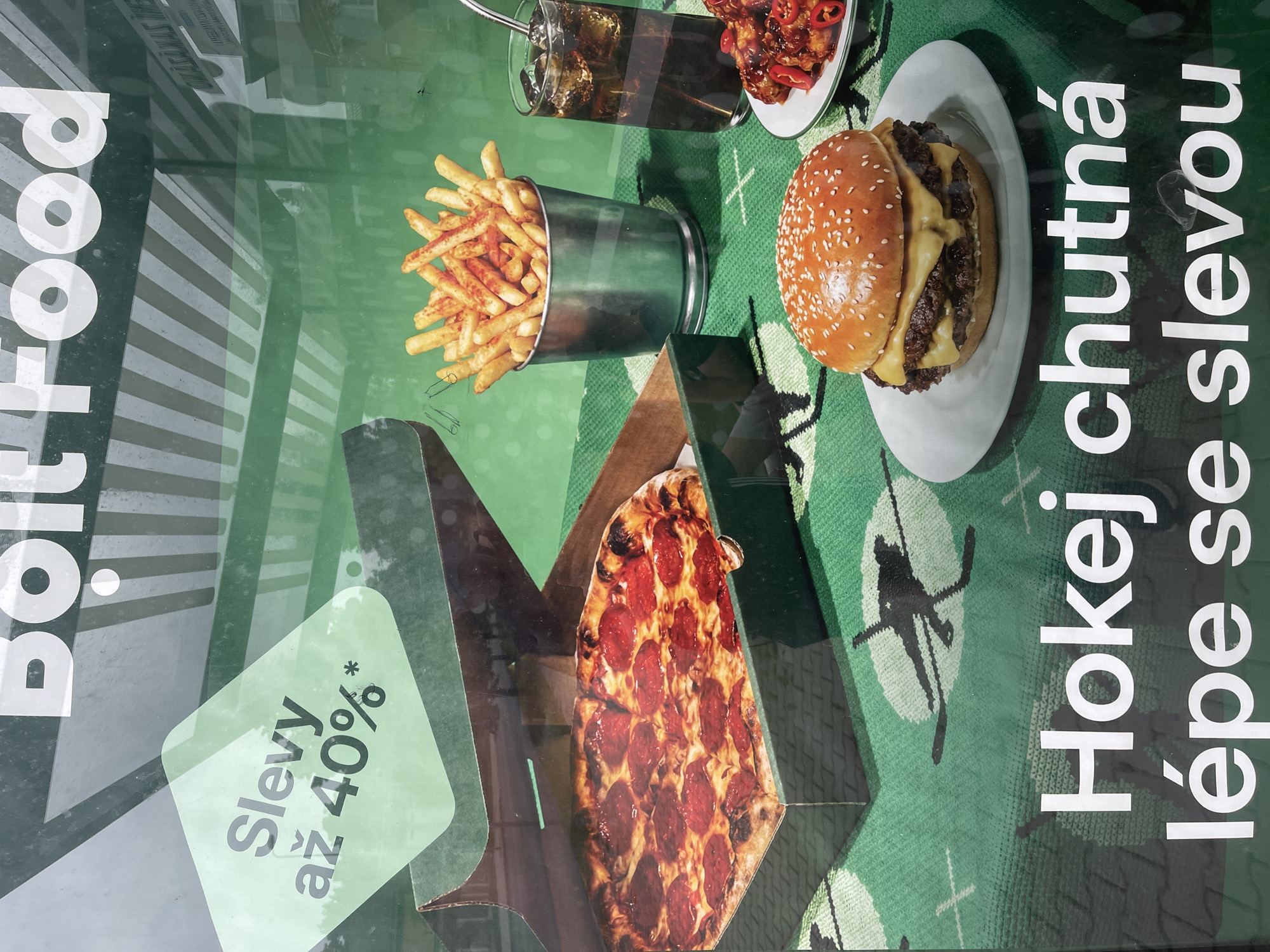 This media file shows a close-up of a promotional display for Bolt Food, a food delivery service. The display features mouth-watering images of food, including a large hamburger, a bucket of french fries, and a pepperoni pizza. These food items are arranged on a green background with a subtle hockey-themed pattern, suggesting a sporty or casual theme for the promotion.
Prominently displayed is the Bolt Food logo and the Czech text "Bolt food," indicating the brand and language. Below the food images, a green sticker or graphic element states "Slevy až 40%*," which translates to "Discounts up to 40%*," highlighting a special offer.
To the right of the food items, large white text is vertically oriented, reading "Hokej chutná lépe se slevou," meaning "Hockey tastes better with a discount." This phrase directly links the food promotion with the hockey theme of the background.
The overall impression is a vibrant and appealing advertisement designed to entice customers to order food through the Bolt Food app, leveraging attractive food imagery and a tempting discount offer. The specific location context of Pilsen, Czechia, is provided, but the image itself does not contain explicit visual cues pointing to this city, other than the Czech language used in the text. The lighting suggests an indoor setting, likely a window display or a poster.