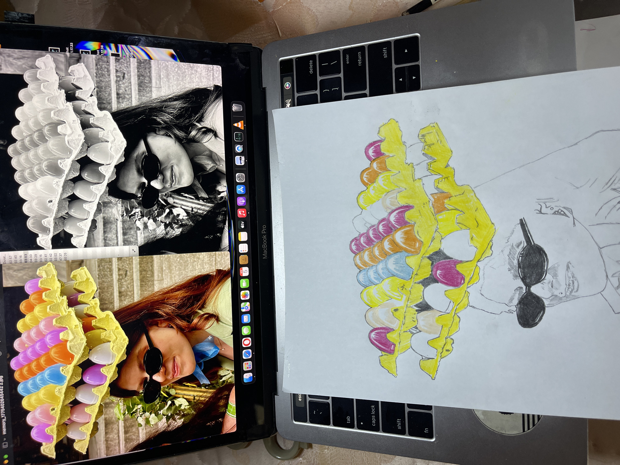 The image shows a desk with a laptop and a drawing. The laptop displays two images: one in color and one in black and white, both showing a woman wearing sunglasses and a crown made of egg cartons filled with plastic eggs. The color image features the crown in various shades of yellow, pink, blue, and orange. The drawing, placed on top of the laptop keyboard, appears to be a work in progress, depicting the same woman and egg carton crown. The drawing is partially colored in, mirroring the color scheme of the image on the laptop screen. The scene is indoors, and the environment is illuminated.