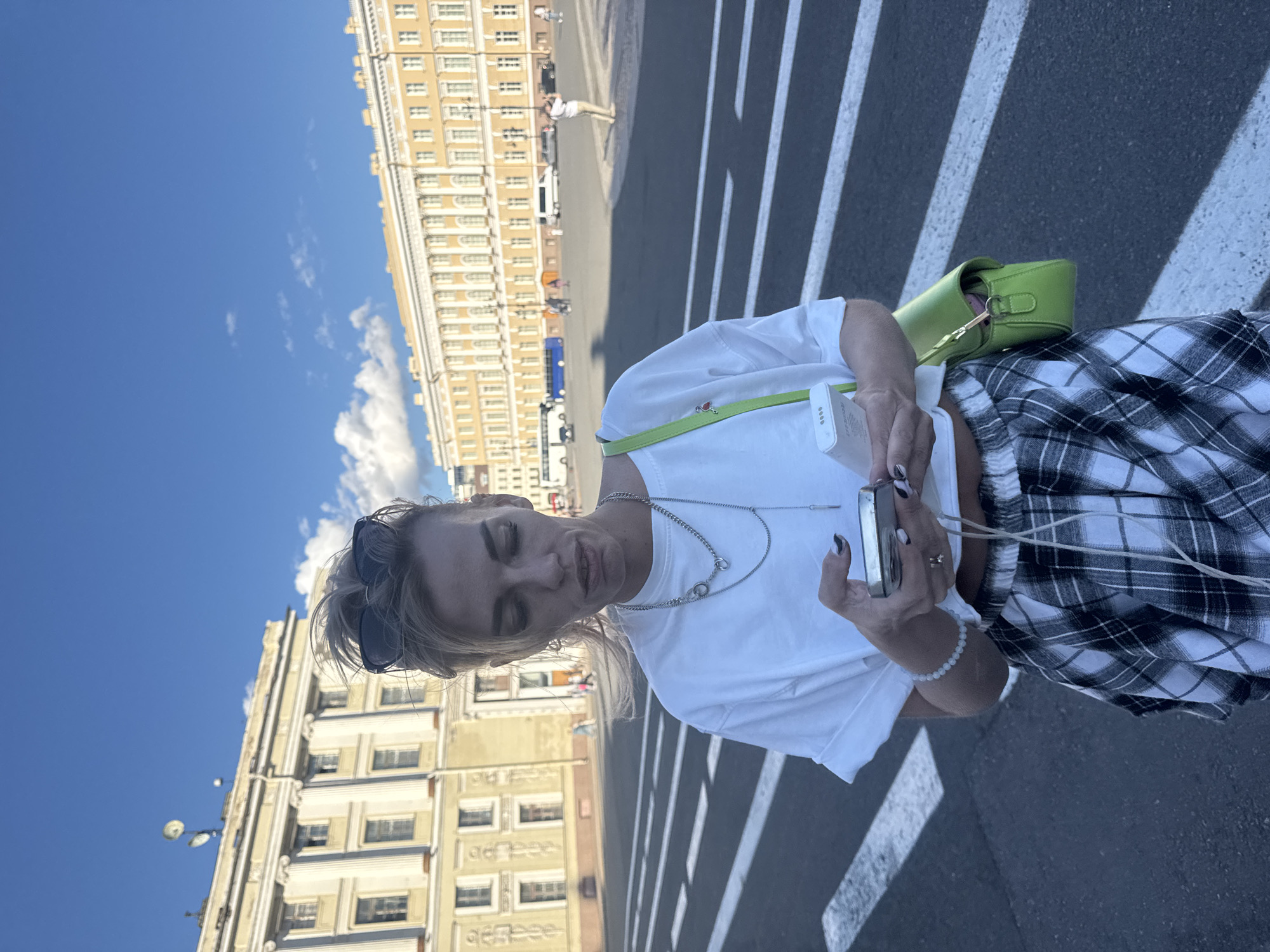 A woman stands on a crosswalk in Saint Petersburg, Russia, in front of a large, ornate building. She is wearing a white t-shirt, a plaid skirt, and sunglasses. She is holding a smartphone and a portable charger. The sky is blue with some clouds. There are buses and cars in the background.