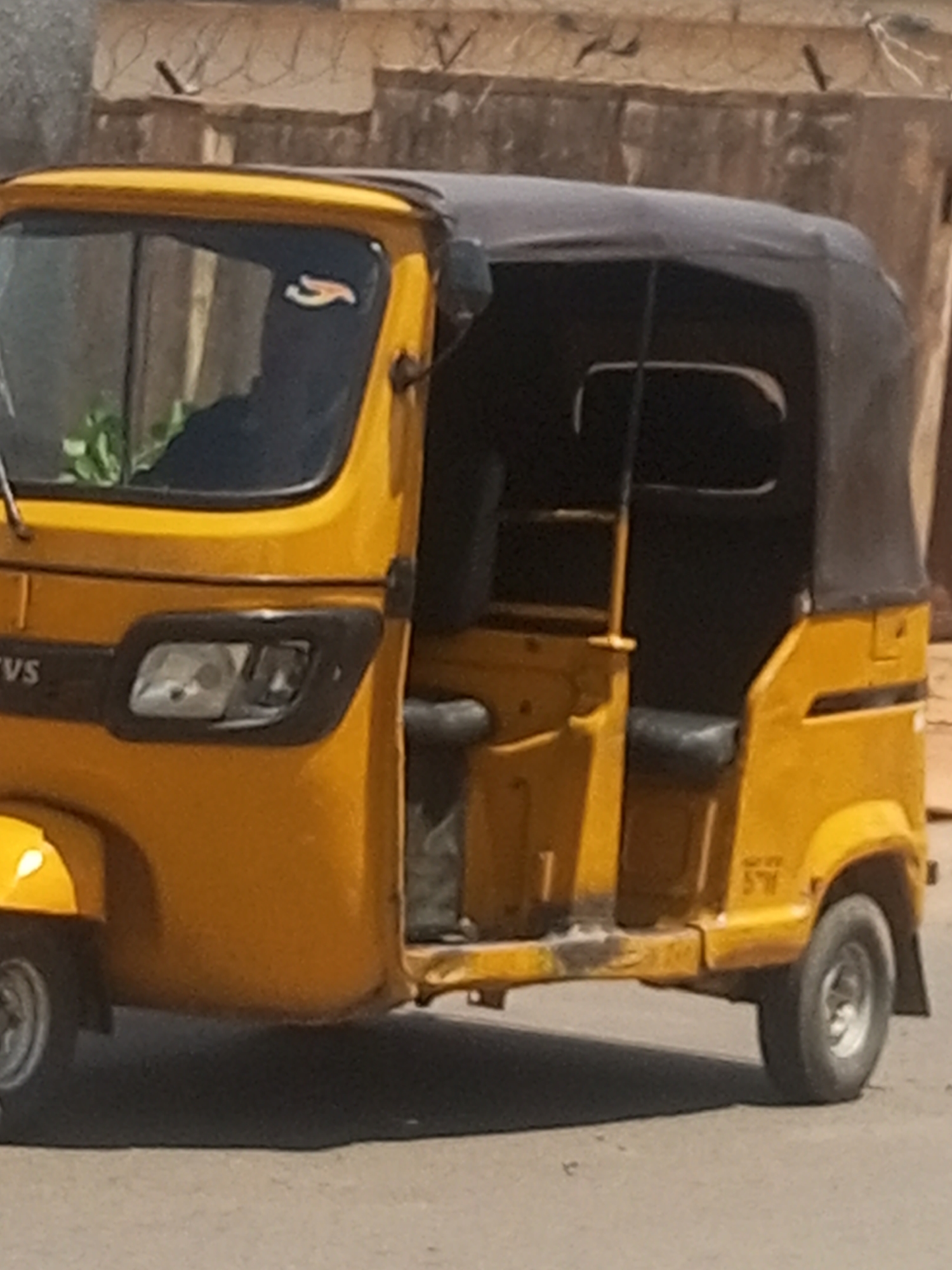 An orange auto-rickshaw with a black canopy is parked on a street. The auto-rickshaw has a single headlight on the left side of the front of the vehicle and a side mirror on the right side. The vehicle has two visible wheels, one in the front and one in the back. The backdrop is a building with a concrete wall and barbed wire at the top. This image was taken in Gombe, Nigeria.