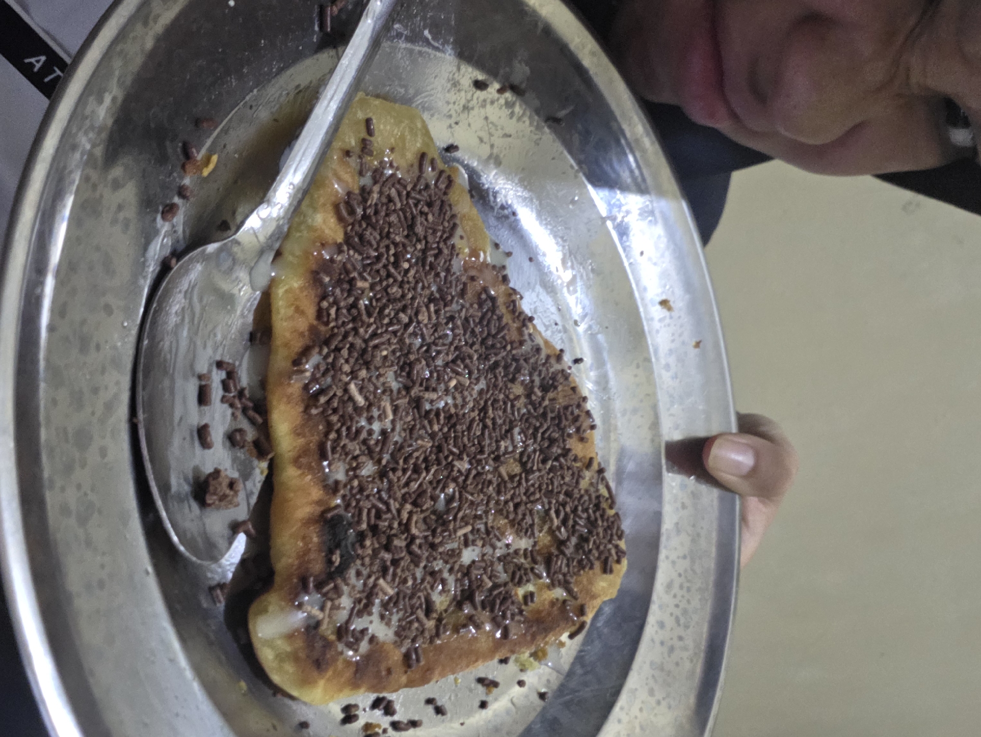 Here is a concise description of the image:
A person is holding a metal plate with a triangle-shaped pancake or crepe covered in chocolate sprinkles and a white sauce. A spoon is on the plate. The background is blurry, but suggests an indoor environment. The location is unknown.