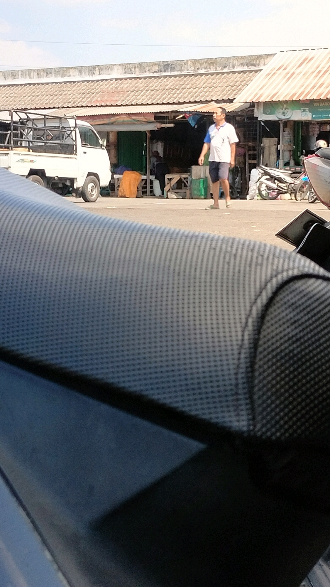 A man is walking in front of a row of market stalls in Wonosari, Indonesia. The man is wearing a white polo shirt with blue trim and dark shorts. He is walking from right to left in the image. Behind him, several market stalls are visible, with awnings and open fronts. A white pickup truck is parked to the left of the stalls. A motorcycle is parked to the right of the stalls. The ground is paved. The sky is clear and blue. The foreground of the image is filled with the dark, textured seat of a motorcycle.